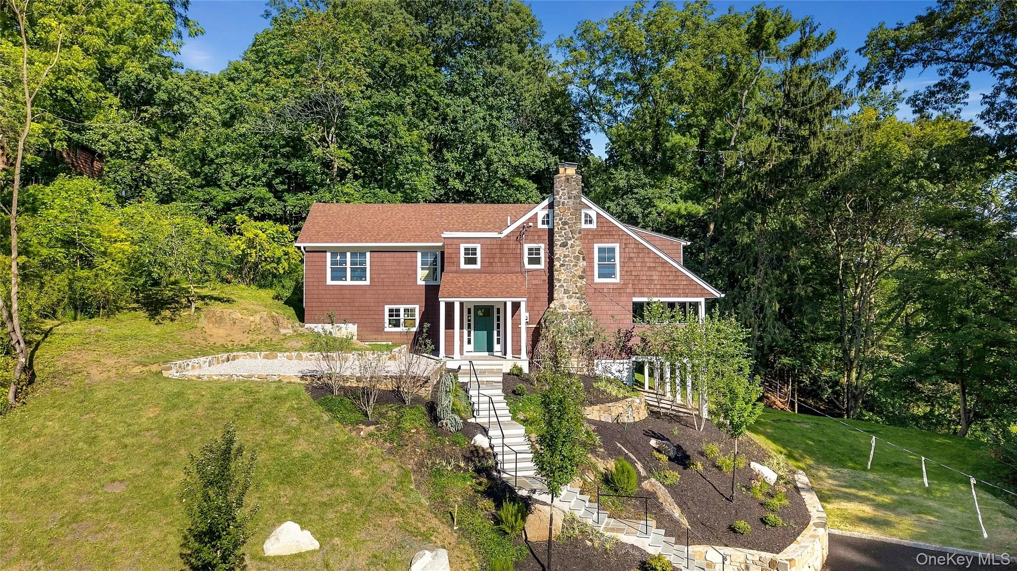 Shingle-sided residence with a stone chimney, a prominent green entry door, and a multi-level sloped landscape with stone retaining walls and steps Shingle-sided residence with a stone chimney, a prominent green entry door, and a multi-level sloped landscape with stone retaining walls and steps