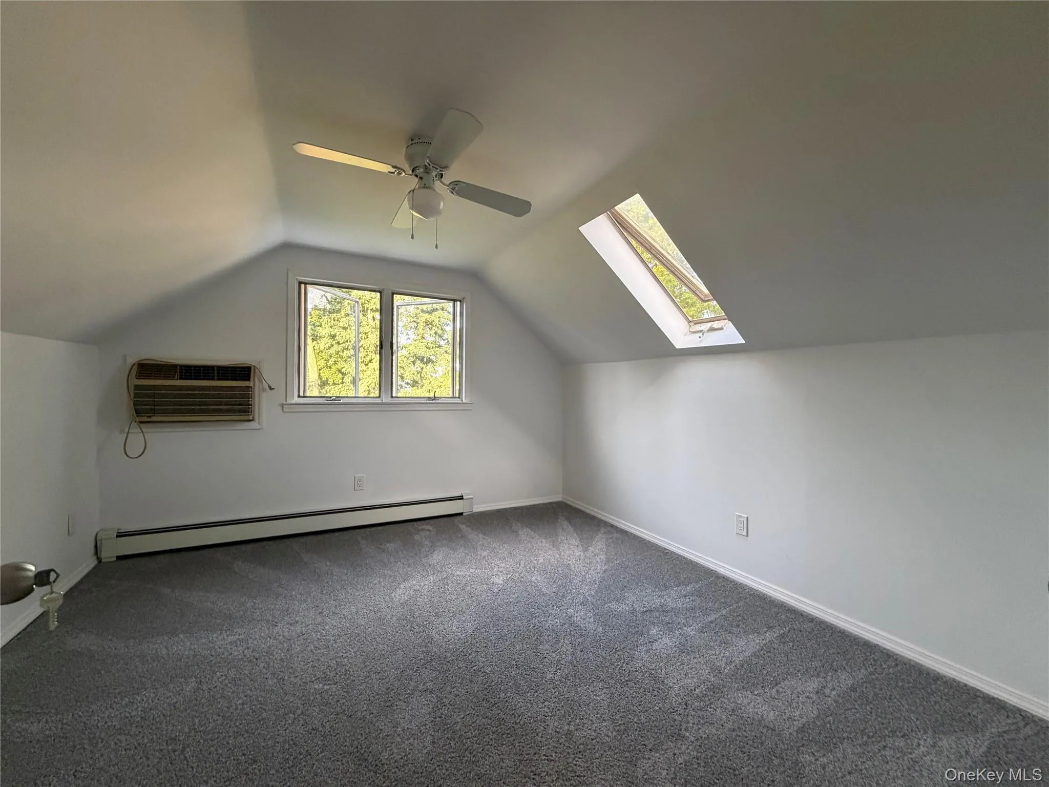 Vaulted ceiling with a ceiling fan, skylight, wall-mounted air conditioning unit, baseboard heating, and gray carpeting Vaulted ceiling with a ceiling fan, skylight, wall-mounted air conditioning unit, baseboard heating, and gray carpeting