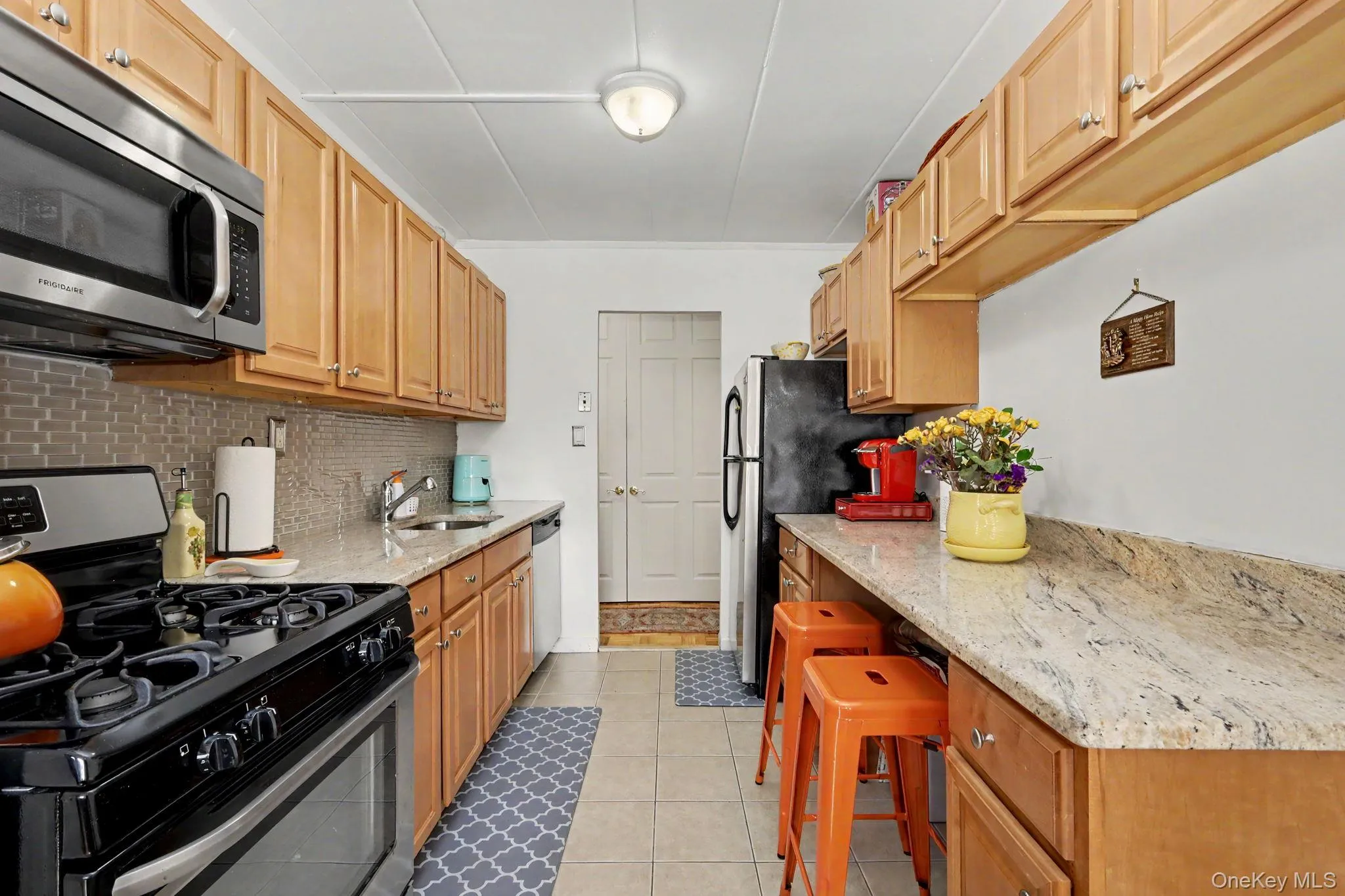 Galley kitchen featuring light wood cabinetry, stone-finish countertops, tile backsplash, stainless steel microwave, and tile flooring Galley kitchen featuring light wood cabinetry, stone-finish countertops, tile backsplash, stainless steel microwave, and tile flooring