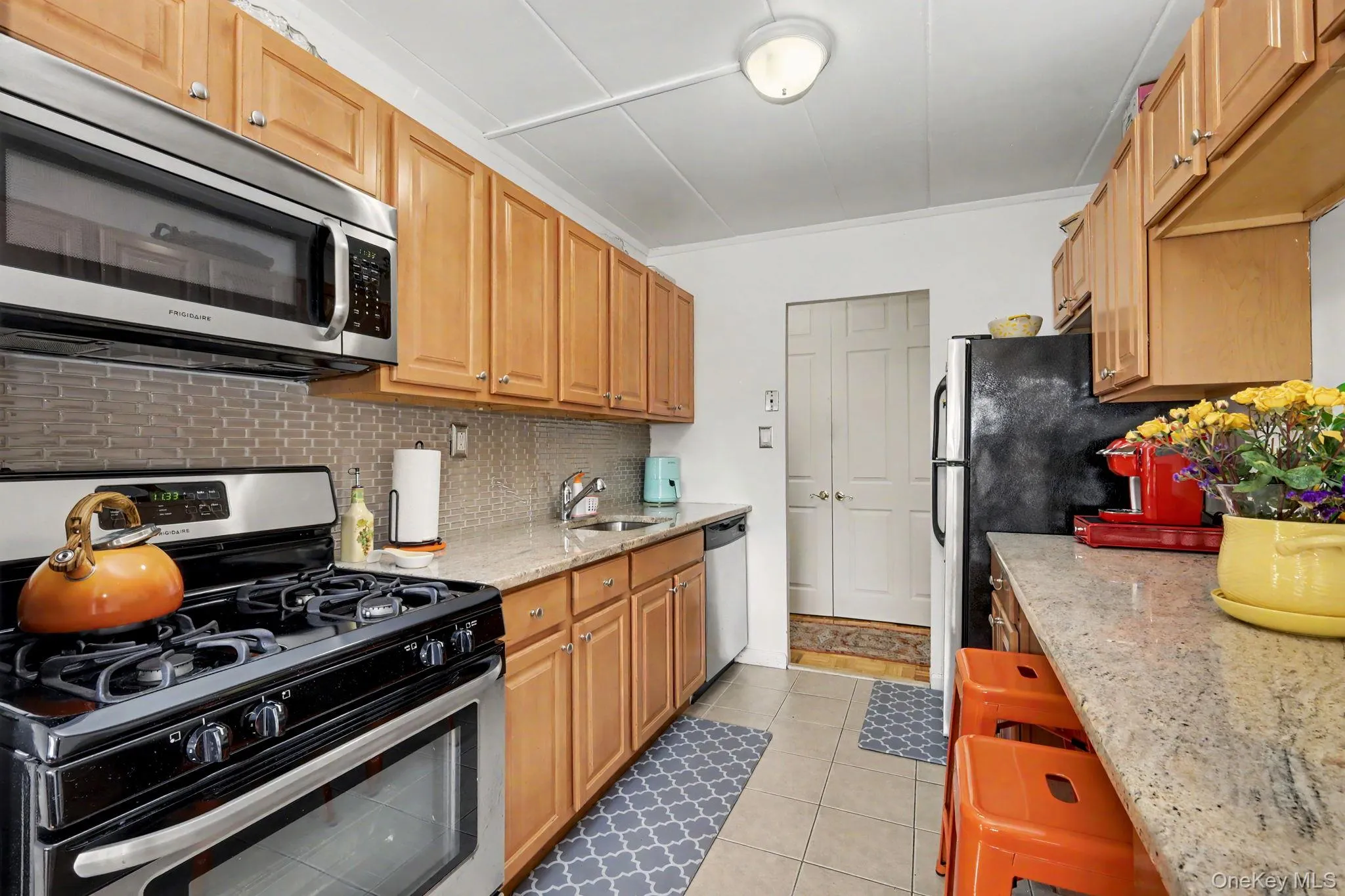 Galley kitchen featuring light wood cabinetry, stone-finish countertops, tile backsplash, stainless steel microwave, and tile flooring Galley kitchen featuring light wood cabinetry, stone-finish countertops, tile backsplash, stainless steel microwave, and tile flooring