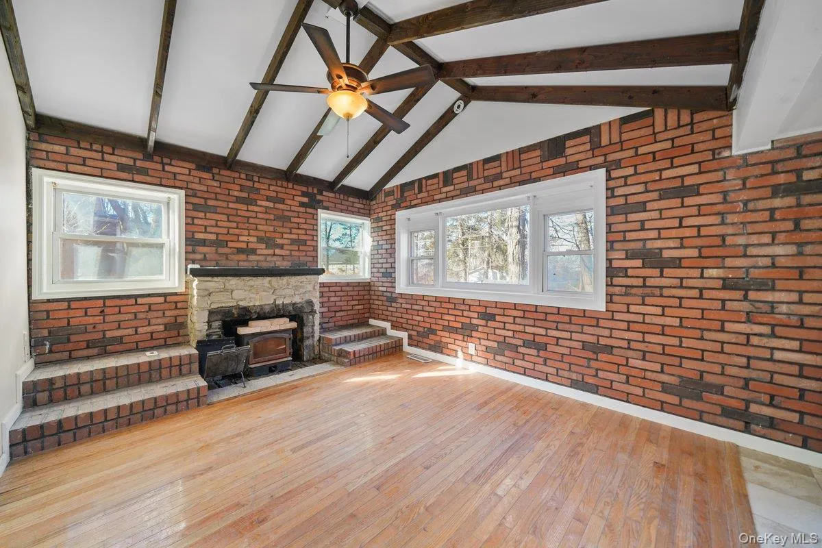 Unfurnished living room featuring brick wall, hardwood / wood-style flooring, a ceiling fan, and a wood stove Unfurnished living room featuring brick wall, hardwood / wood-style flooring, a ceiling fan, and a wood stove