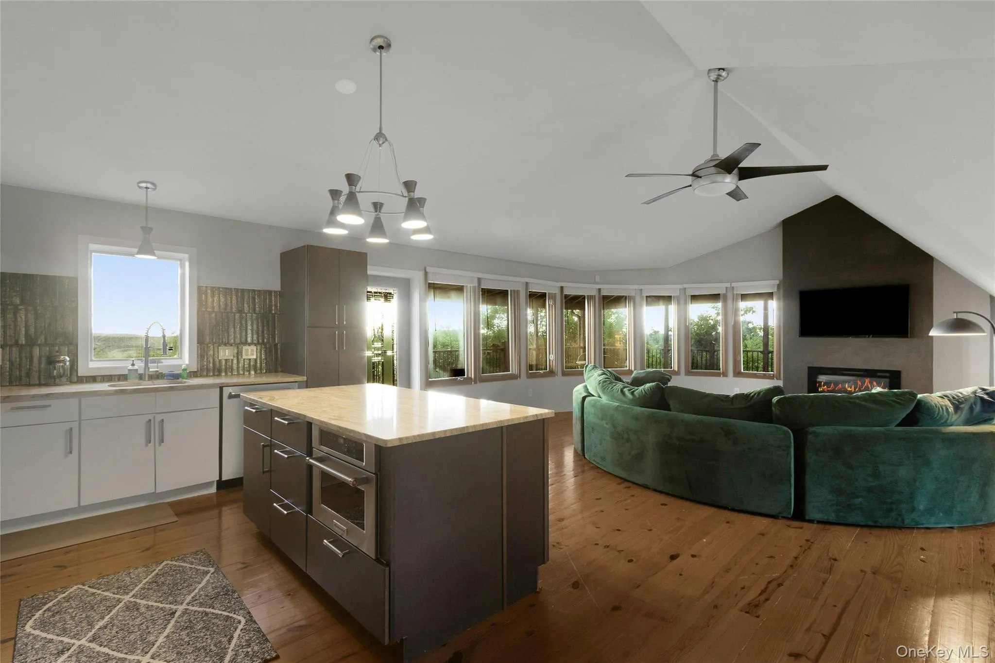 Open-concept living area featuring wood-finish flooring, a kitchen island with built-in microwave, an integrated fireplace, and a vaulted ceiling with a ceiling fan Open-concept living area featuring wood-finish flooring, a kitchen island with built-in microwave, an integrated fireplace, and a vaulted ceiling with a ceiling fan