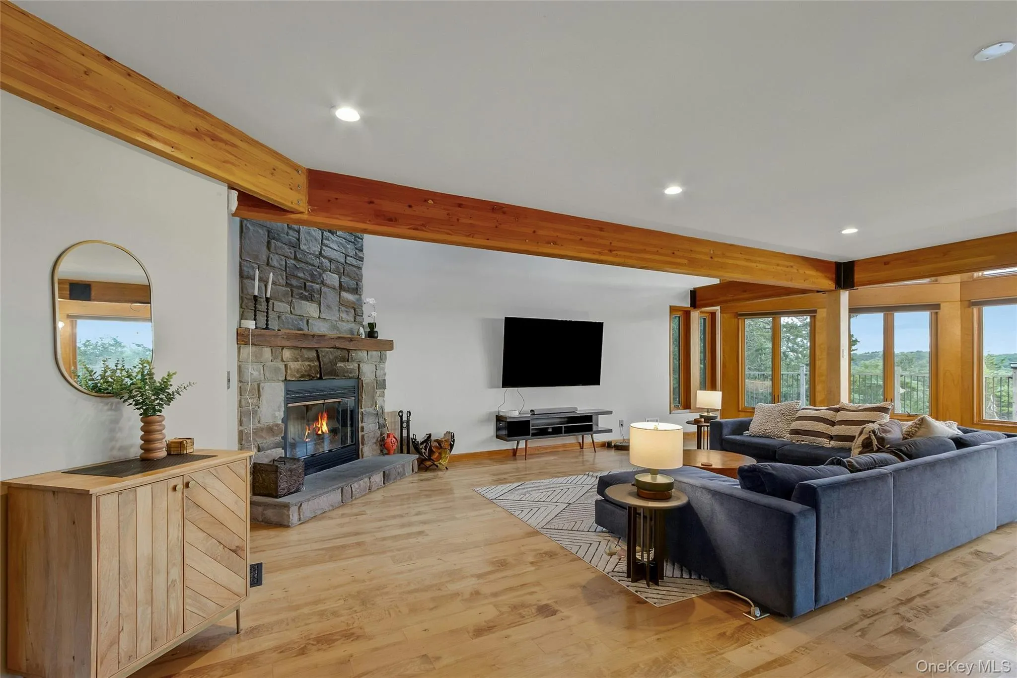 Stone-clad fireplace with hearth, wood-finish flooring, exposed wood beams, recessed lighting, and multiple windows Stone-clad fireplace with hearth, wood-finish flooring, exposed wood beams, recessed lighting, and multiple windows