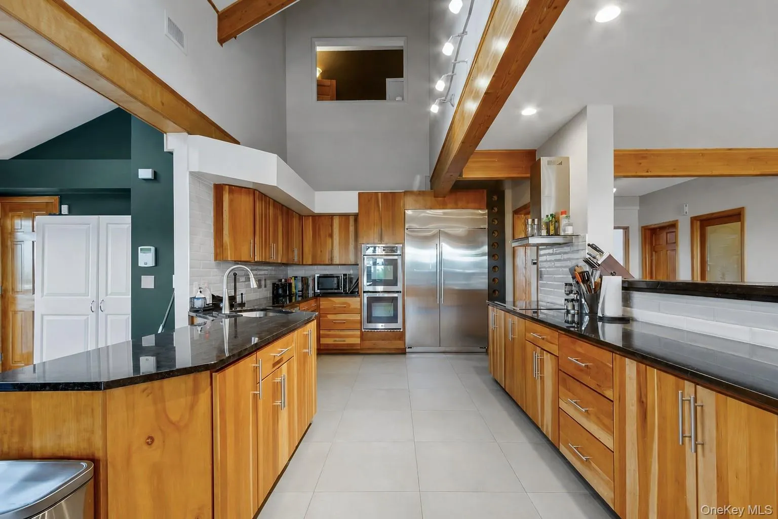 Kitchen featuring natural wood cabinetry, dark countertops, a stainless steel refrigerator, double ovens, and a tile backsplash Kitchen featuring natural wood cabinetry, dark countertops, a stainless steel refrigerator, double ovens, and a tile backsplash