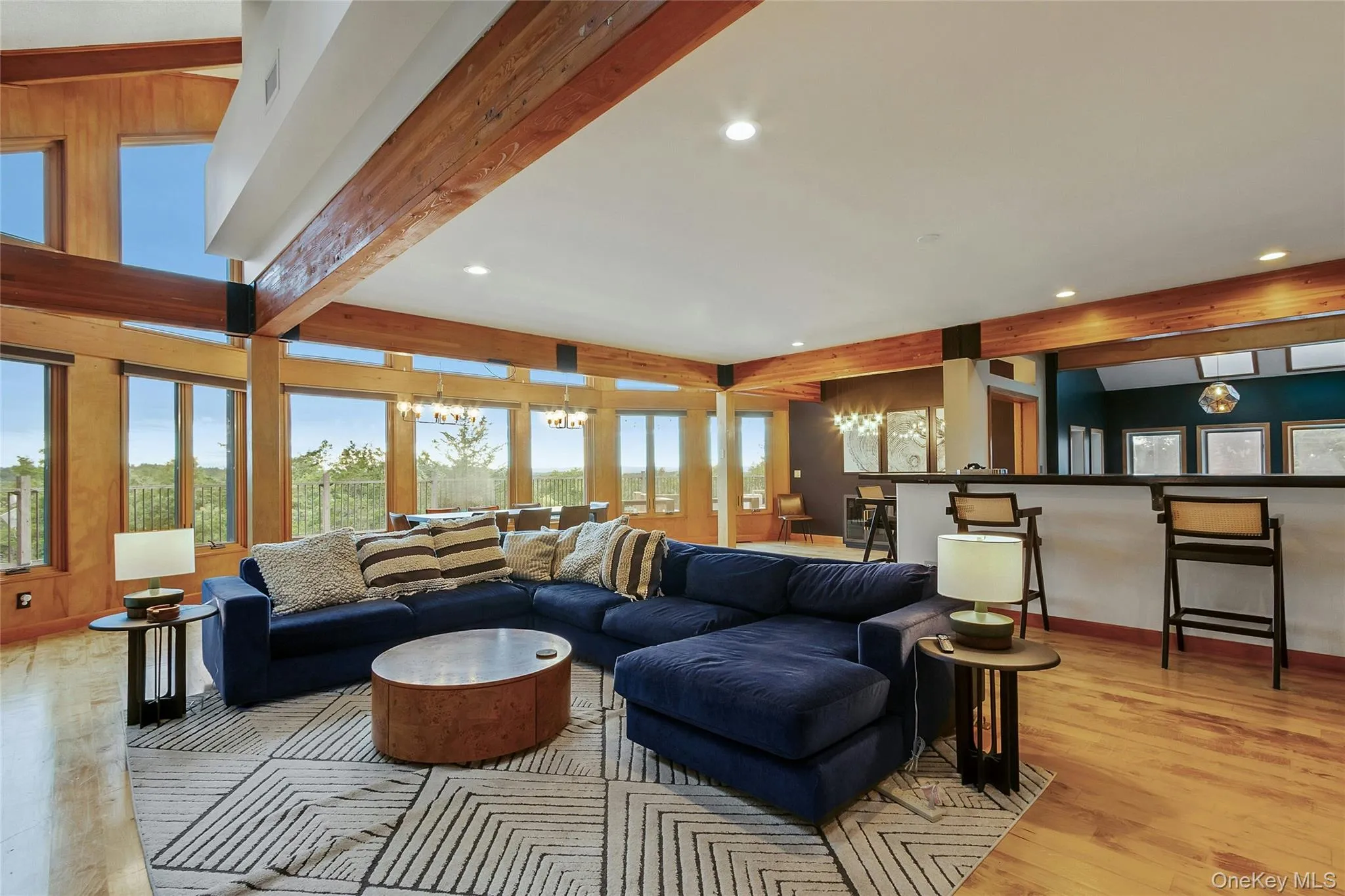 Expansive living space featuring exposed wood beams, wood-finish flooring, a full-height window wall, recessed lighting, and a built-in bar with counter seating Expansive living space featuring exposed wood beams, wood-finish flooring, a full-height window wall, recessed lighting, and a built-in bar with counter seating