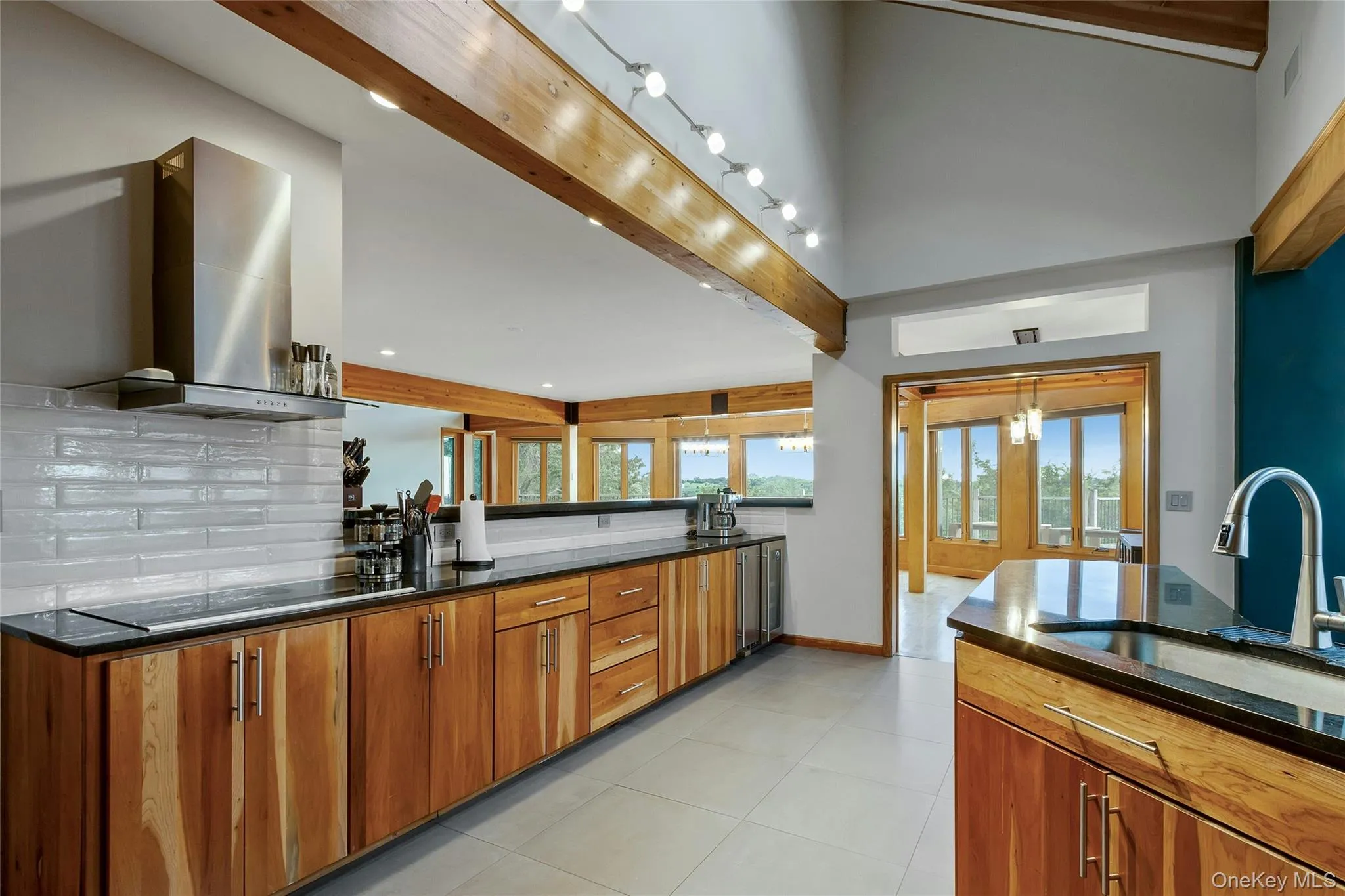 Vaulted ceiling with exposed wood beams, stainless steel range hood, natural wood cabinetry, black countertops, and a white subway tile backsplash Vaulted ceiling with exposed wood beams, stainless steel range hood, natural wood cabinetry, black countertops, and a white subway tile backsplash
