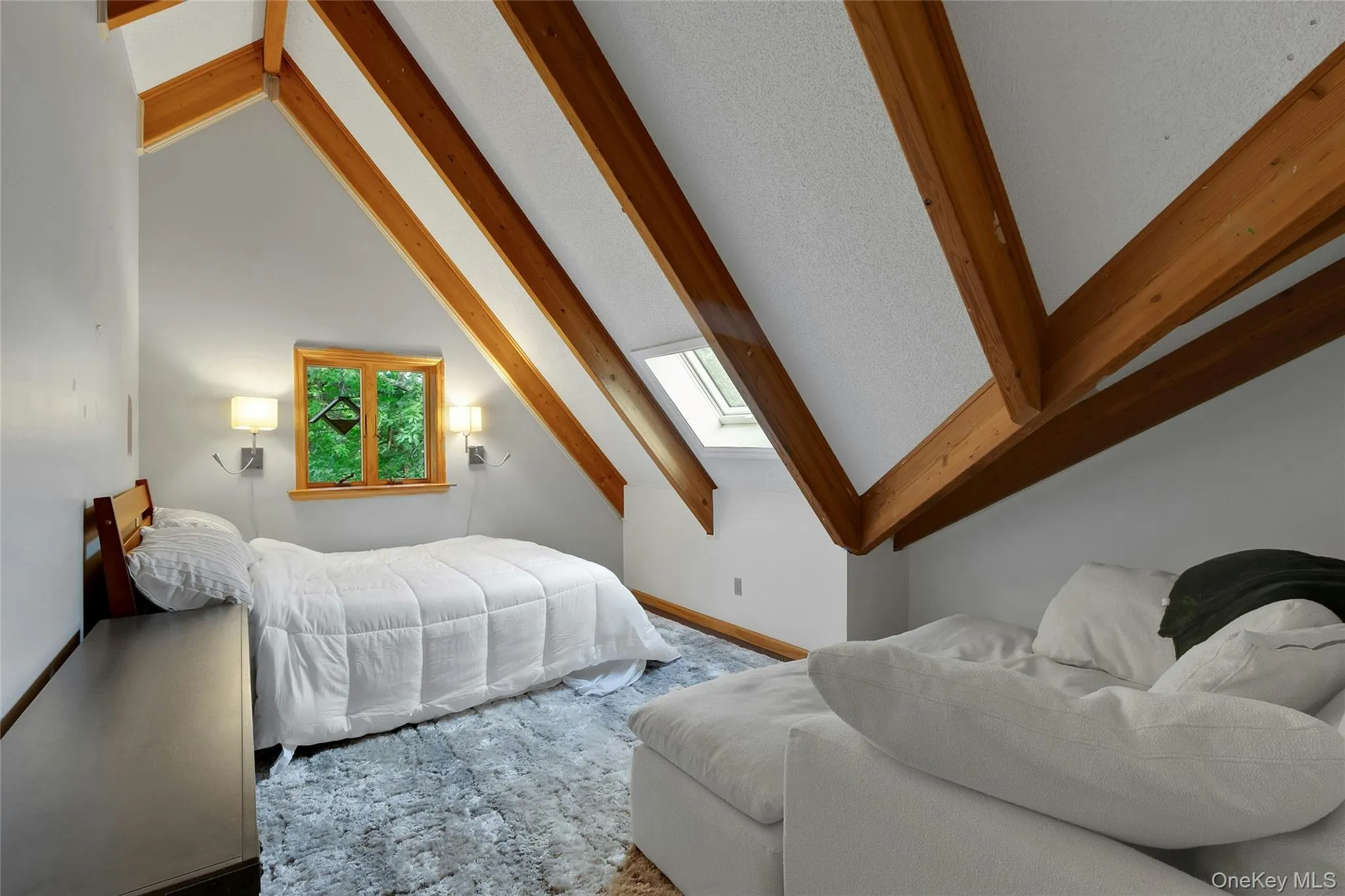 Vaulted ceiling with exposed wood beams, wood-frame window, two wall-mounted sconces, and a skylight Vaulted ceiling with exposed wood beams, wood-frame window, two wall-mounted sconces, and a skylight