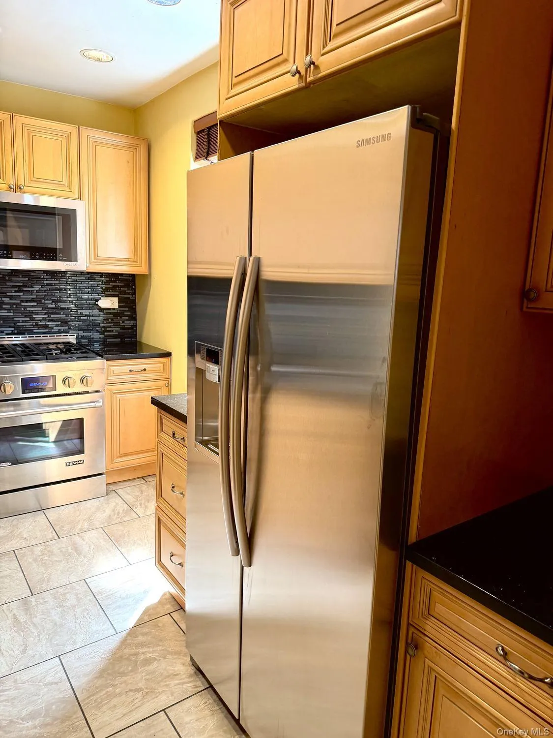 Wood-finish cabinetry, dark countertops, tiled backsplash, stainless steel appliances, and tiled flooring Wood-finish cabinetry, dark countertops, tiled backsplash, stainless steel appliances, and tiled flooring