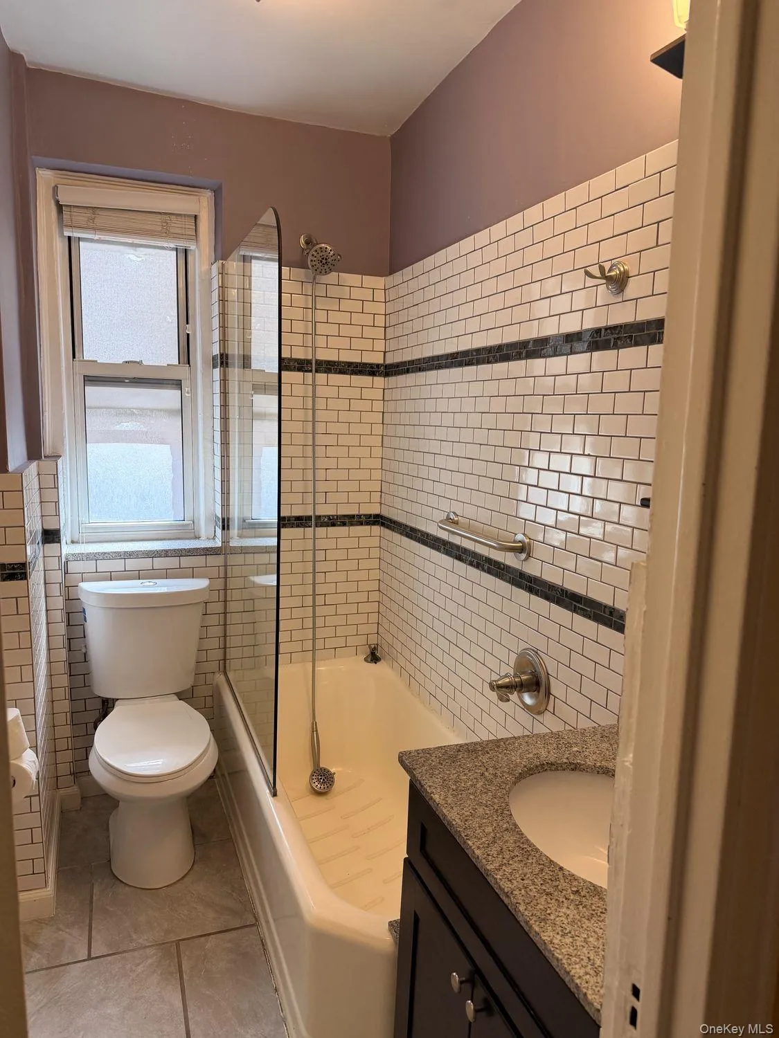 White subway tile surround with a dark accent band, built-in bathtub with glass partition, and a vanity featuring a granite-style countertop and integrated sink White subway tile surround with a dark accent band, built-in bathtub with glass partition, and a vanity featuring a granite-style countertop and integrated sink