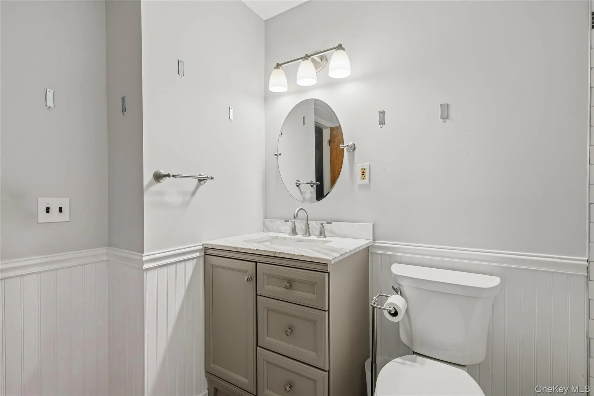 Vanity with a light-toned countertop, beadboard wainscoting, wall-mounted lighting fixture, and an oval mirror Vanity with a light-toned countertop, beadboard wainscoting, wall-mounted lighting fixture, and an oval mirror