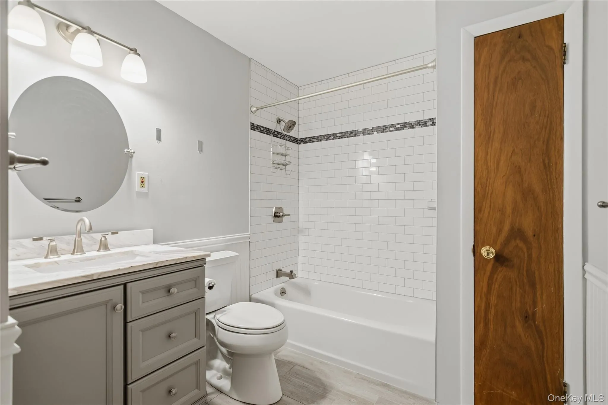 White subway tile shower surround with mosaic accent, gray vanity with stone countertop, round wall mirror, and wood-finish door White subway tile shower surround with mosaic accent, gray vanity with stone countertop, round wall mirror, and wood-finish door