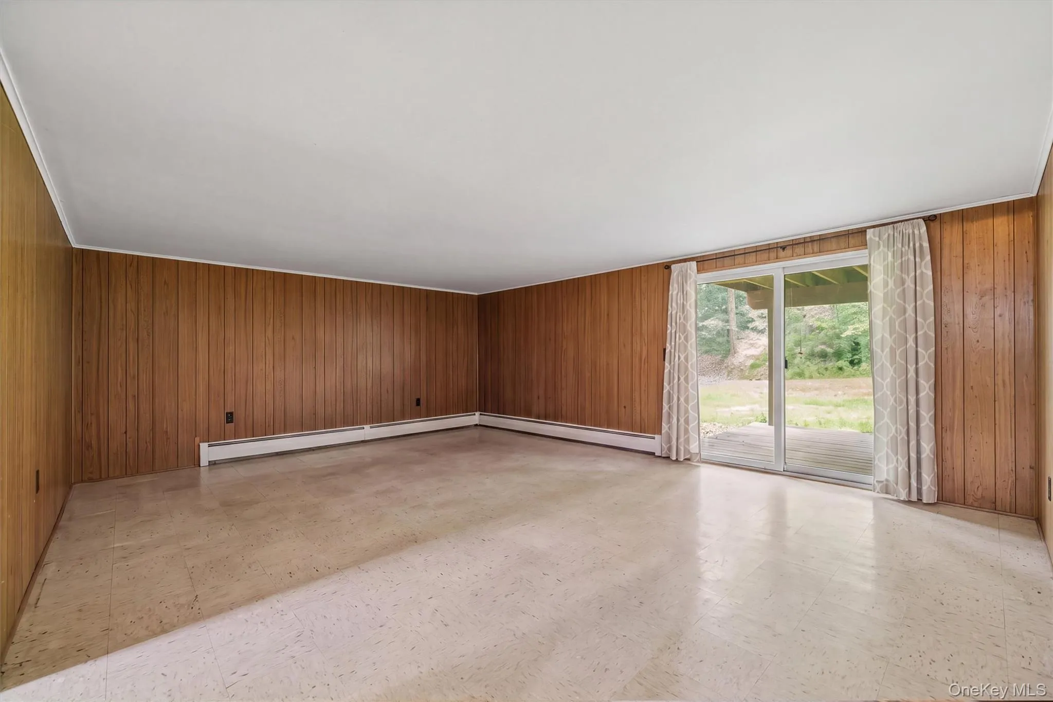 Spacious room featuring wood paneling, light-toned flooring, and baseboard heating Spacious room featuring wood paneling, light-toned flooring, and baseboard heating