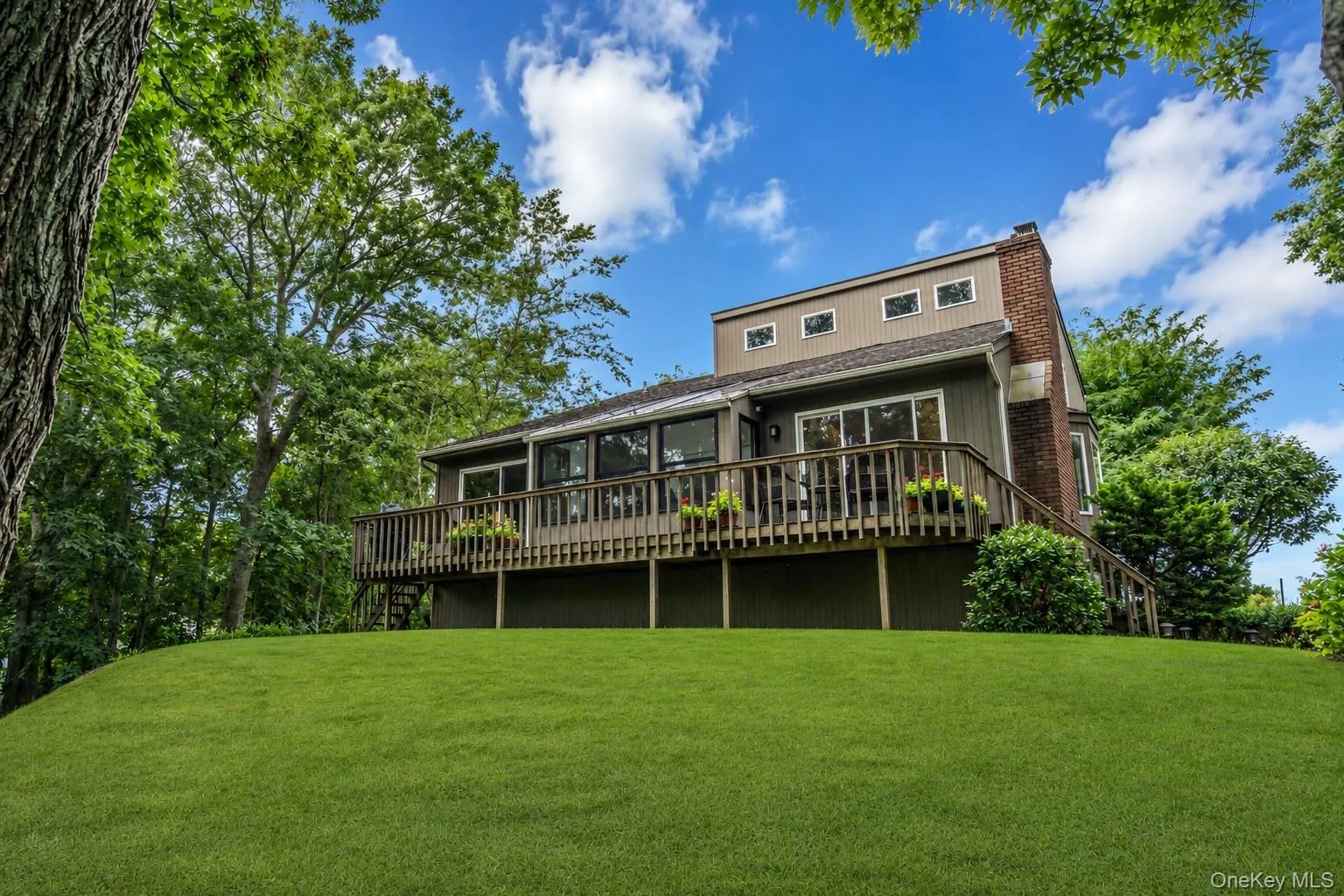 Elevated residence featuring a broad wooden deck, a brick chimney, multiple large windows, and an expansive lawn Elevated residence featuring a broad wooden deck, a brick chimney, multiple large windows, and an expansive lawn