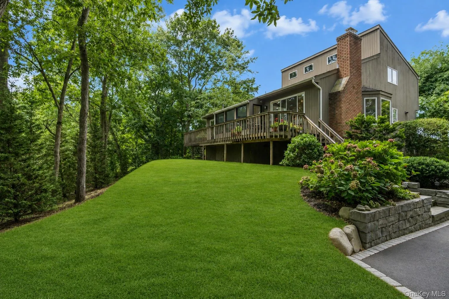 Elevated residence featuring a broad wooden deck, a brick chimney, multiple large windows, and a spacious yard Elevated residence featuring a broad wooden deck, a brick chimney, multiple large windows, and a spacious yard