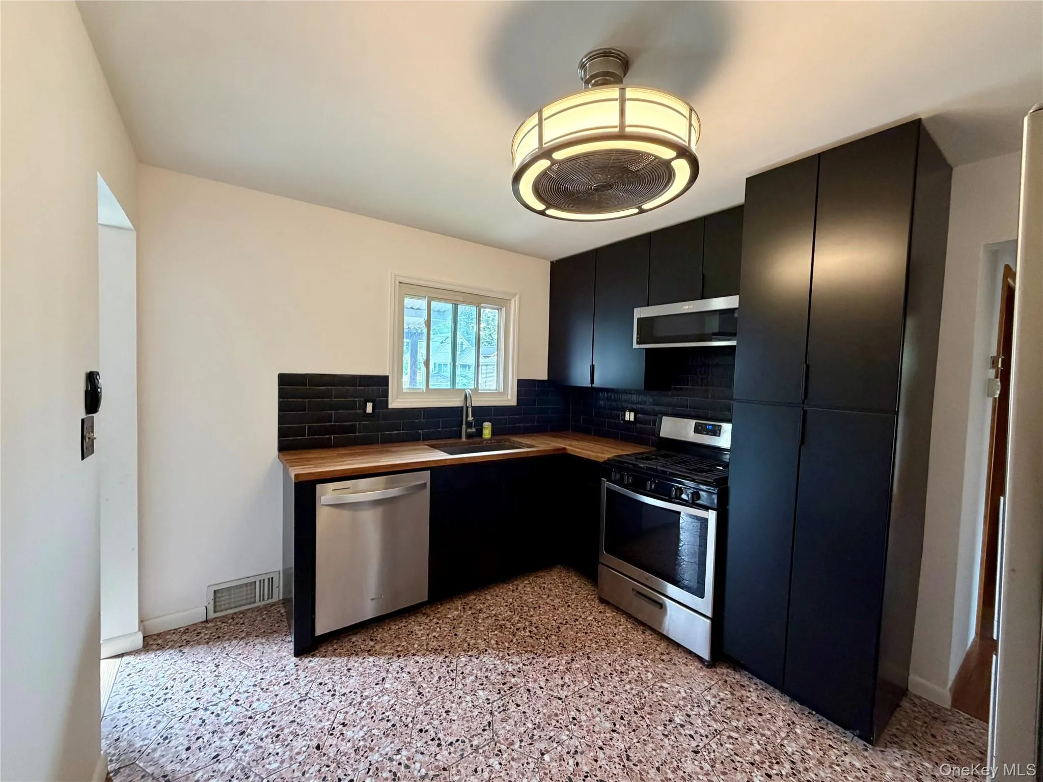 Kitchen featuring black cabinetry, wood-finish countertops, stainless steel appliances, a black subway tile backsplash, and composite flooring Kitchen featuring black cabinetry, wood-finish countertops, stainless steel appliances, a black subway tile backsplash, and composite flooring