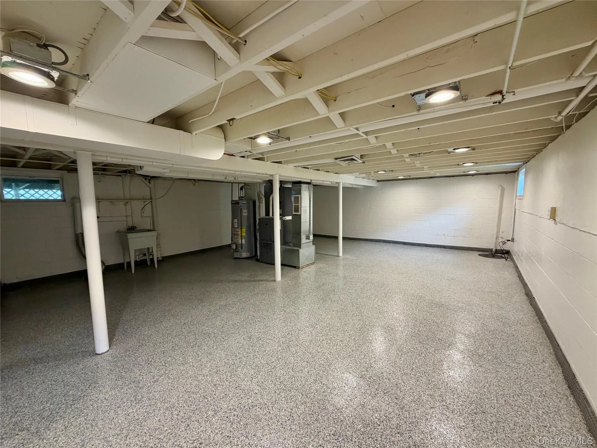 Spacious basement with exposed ceiling beams, a utility sink, and wall-mounted lighting Spacious basement with exposed ceiling beams, a utility sink, and wall-mounted lighting