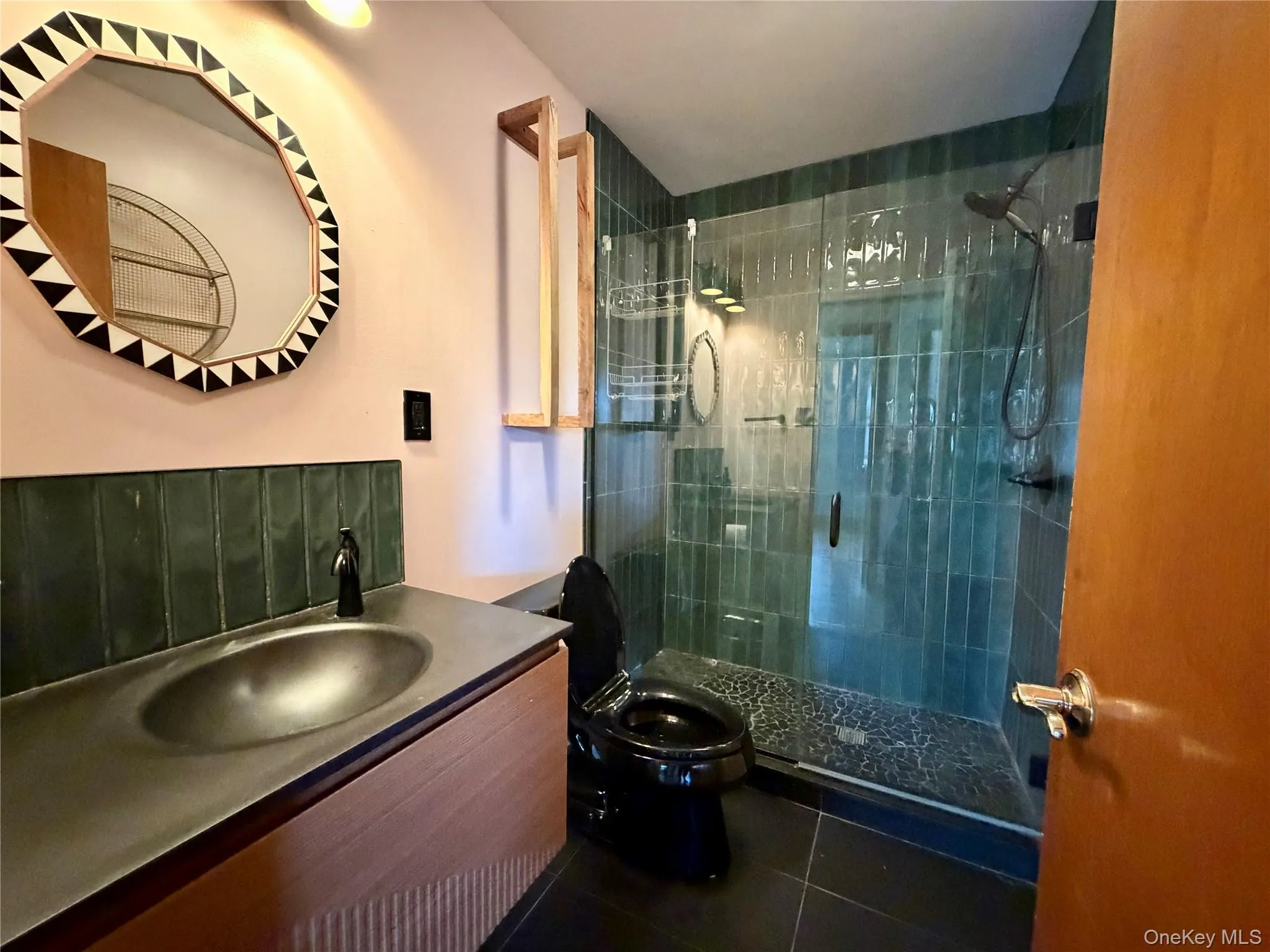 Vanity with an integrated basin and dark countertop, glass-enclosed shower with subway tile surround, dark toilet, and patterned decorative mirror Vanity with an integrated basin and dark countertop, glass-enclosed shower with subway tile surround, dark toilet, and patterned decorative mirror