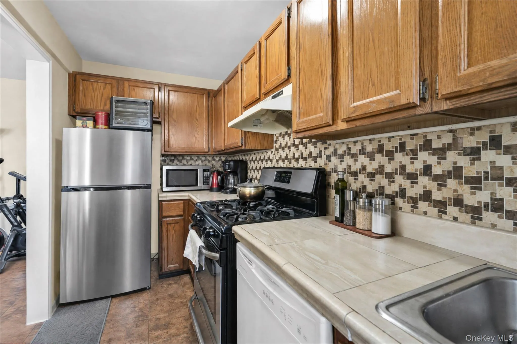 Wood-finish cabinetry, stainless steel refrigerator, patterned tile backsplash, integrated stainless steel sink, and light-toned countertops Wood-finish cabinetry, stainless steel refrigerator, patterned tile backsplash, integrated stainless steel sink, and light-toned countertops