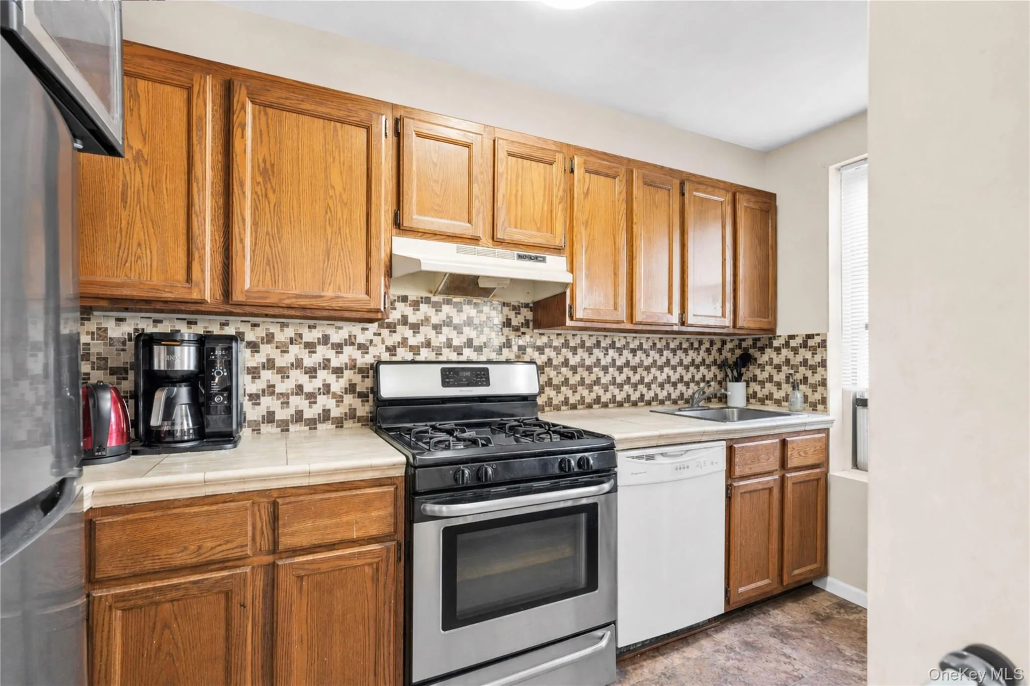 Kitchen featuring wood cabinetry, stainless steel gas range, white dishwasher, tile countertops, and a mosaic tile backsplash Kitchen featuring wood cabinetry, stainless steel gas range, white dishwasher, tile countertops, and a mosaic tile backsplash