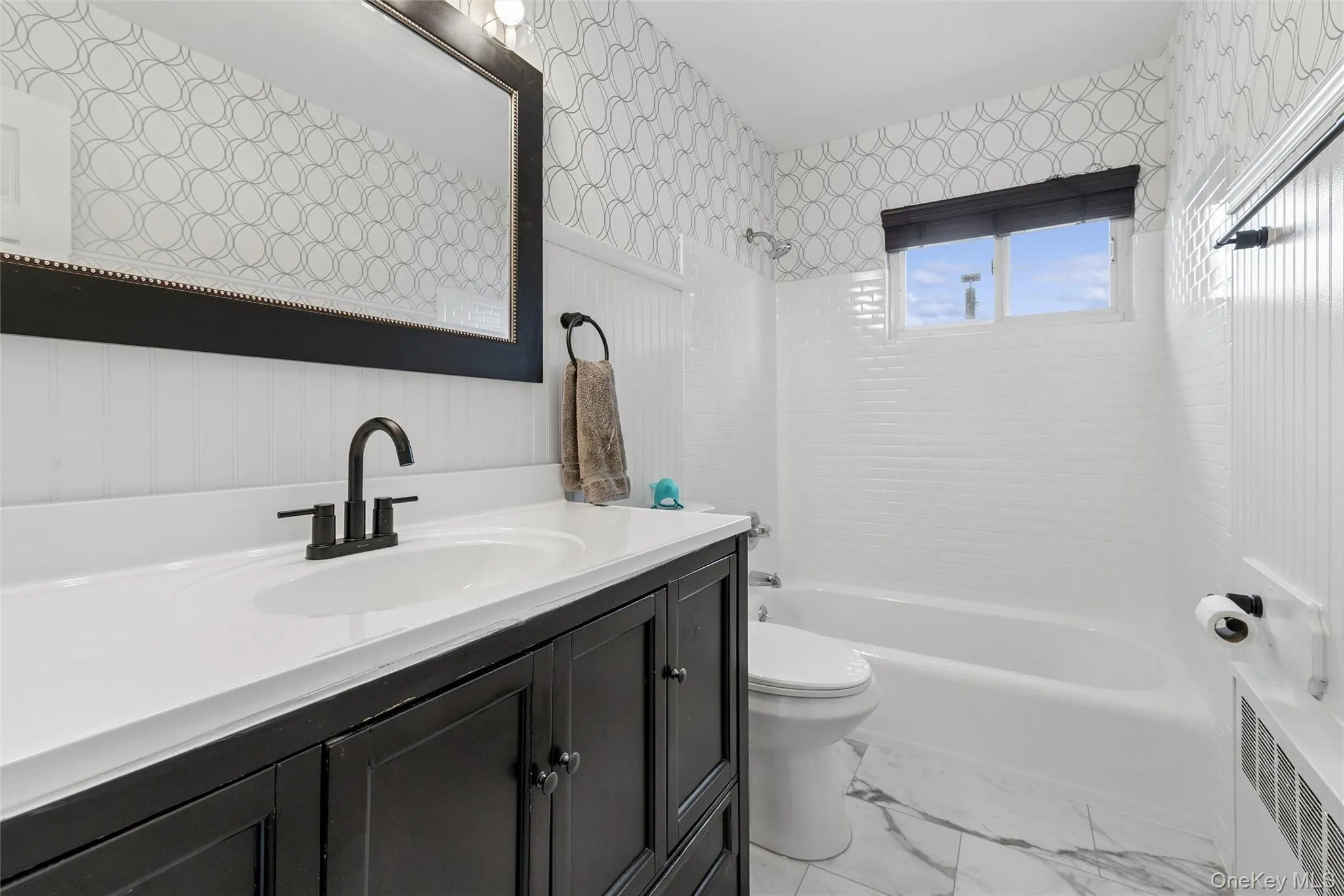 Bathroom featuring a dark wood-finish vanity with a white countertop, matte black fixtures, an undermount sink, a framed mirror, and patterned wallpaper Bathroom featuring a dark wood-finish vanity with a white countertop, matte black fixtures, an undermount sink, a framed mirror, and patterned wallpaper