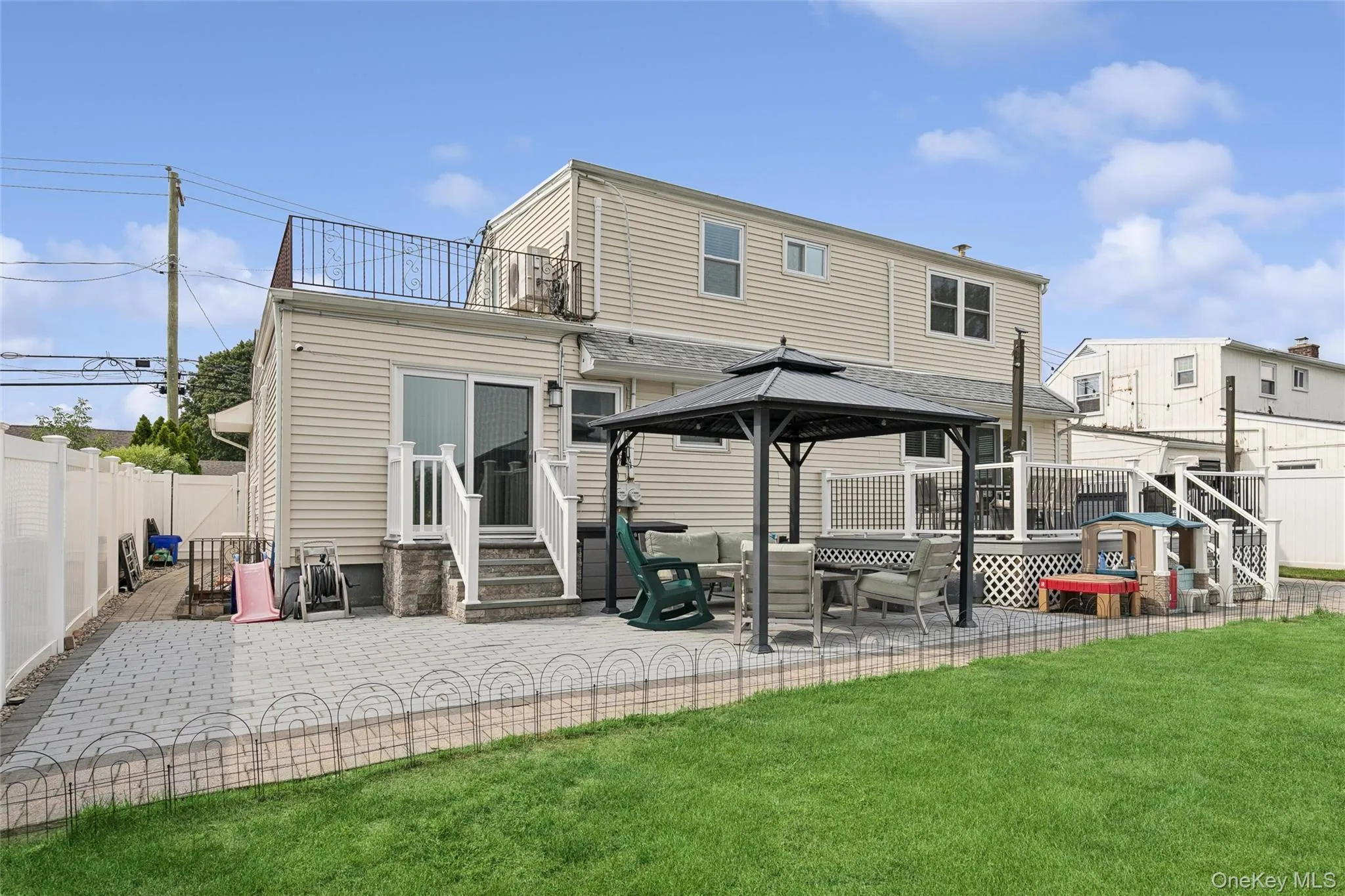 Expansive backyard featuring a paver patio, dedicated lawn space, an elevated deck with white railing, and a private second-story balcony with decorative metalwork Expansive backyard featuring a paver patio, dedicated lawn space, an elevated deck with white railing, and a private second-story balcony with decorative metalwork