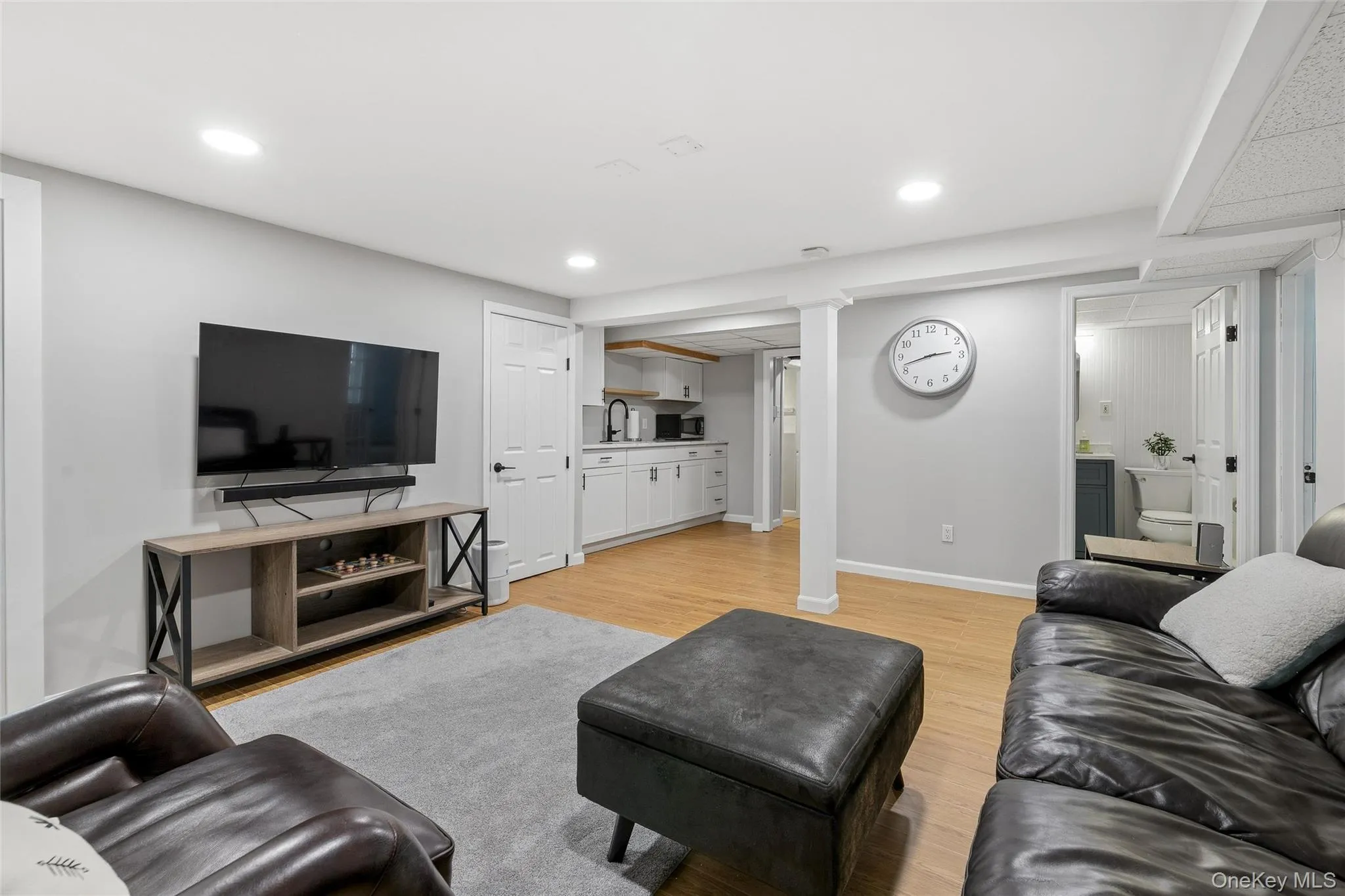 Recessed lighting illuminates this versatile space featuring wood-finish flooring, a contemporary kitchenette with white cabinetry, and a full bathroom with beadboard paneling Recessed lighting illuminates this versatile space featuring wood-finish flooring, a contemporary kitchenette with white cabinetry, and a full bathroom with beadboard paneling