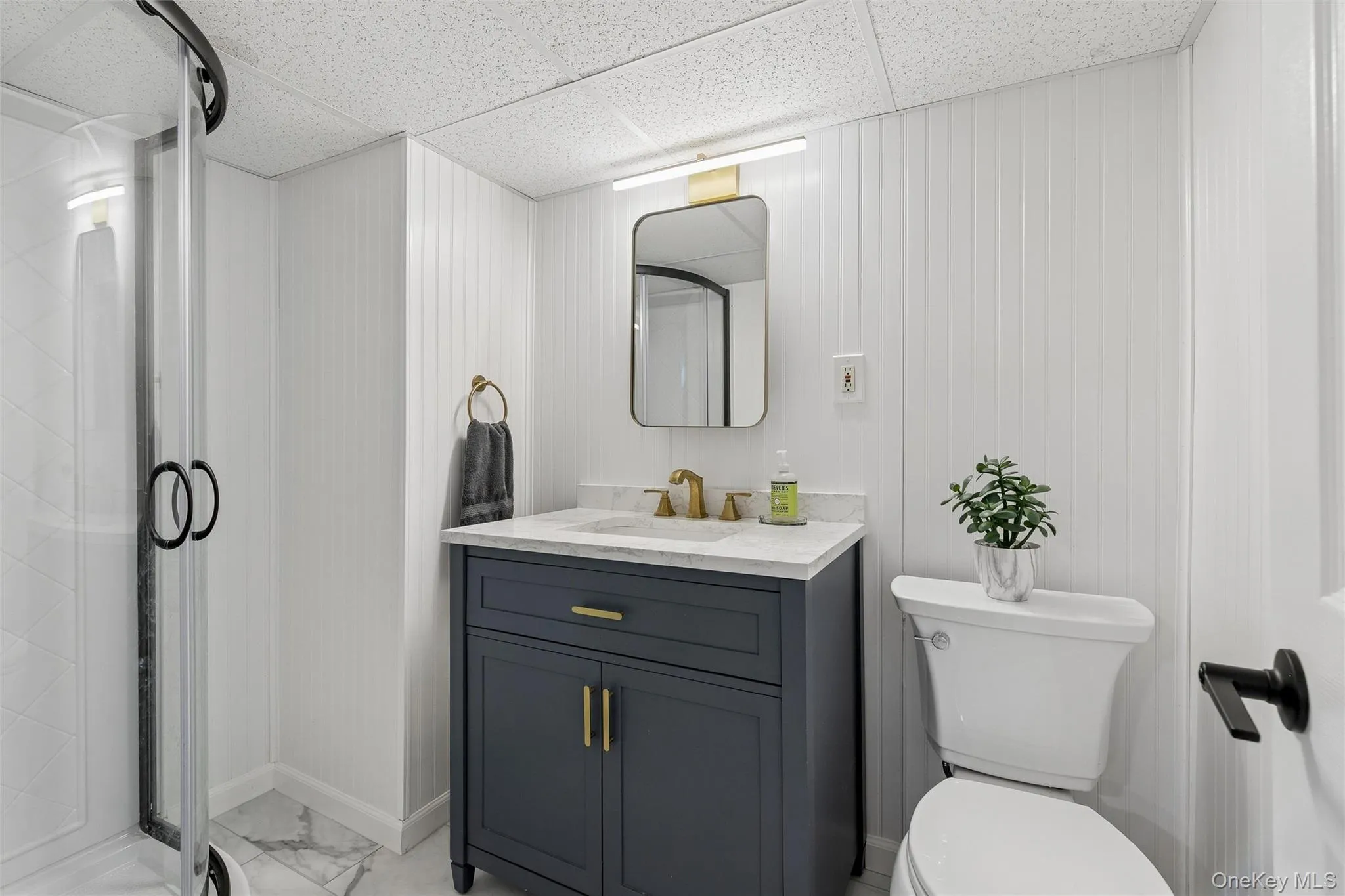 Bathroom with a dark blue vanity, gold-tone fixtures, a glass-enclosed shower, white beadboard paneling, and marble-look floor tiling Bathroom with a dark blue vanity, gold-tone fixtures, a glass-enclosed shower, white beadboard paneling, and marble-look floor tiling