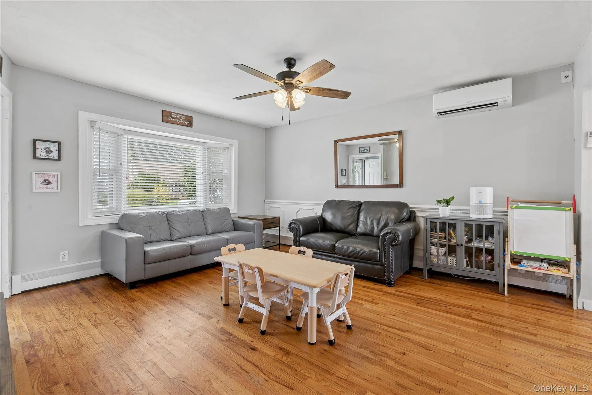 Hardwood-finish flooring, bay window with blinds, ceiling fan, wall-mounted air conditioning unit, and white wainscoting Hardwood-finish flooring, bay window with blinds, ceiling fan, wall-mounted air conditioning unit, and white wainscoting