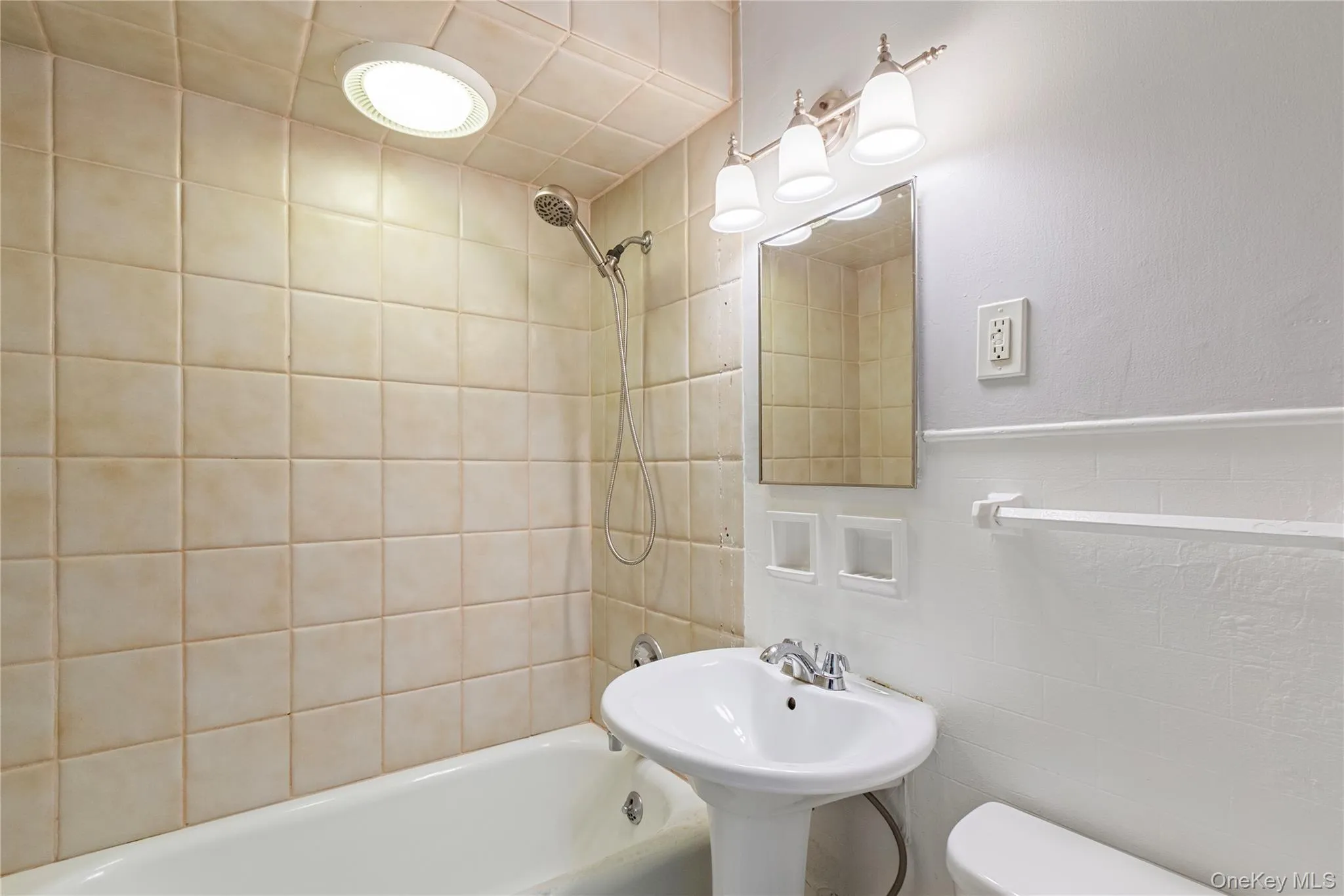 Pedestal sink with chrome fixtures, tiled shower/tub combination, wall-mounted mirror, and vanity lighting Pedestal sink with chrome fixtures, tiled shower/tub combination, wall-mounted mirror, and vanity lighting