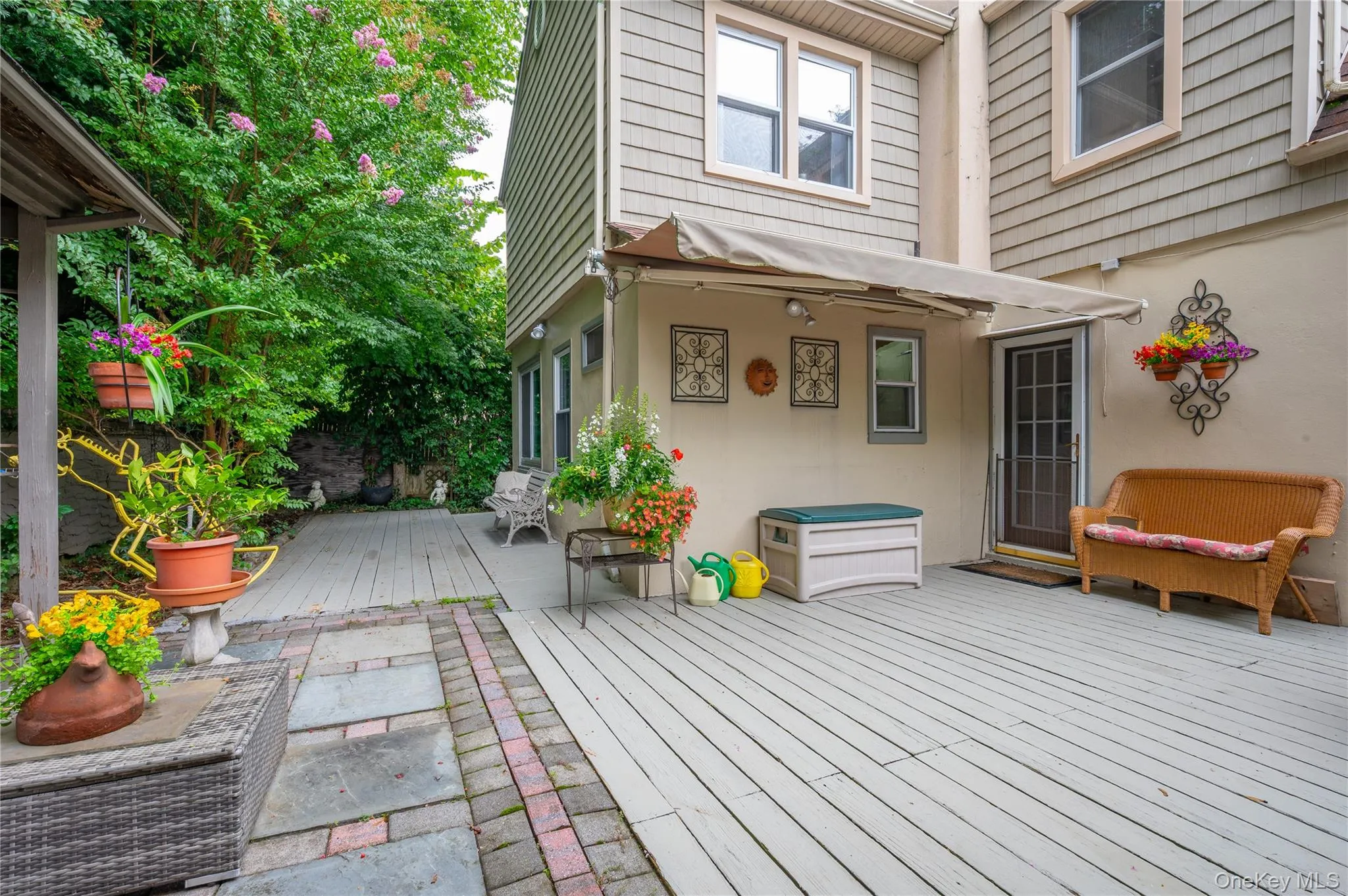 Expansive outdoor deck with a retractable awning, surrounded by established greenery, featuring a separate stone and paver patio area Expansive outdoor deck with a retractable awning, surrounded by established greenery, featuring a separate stone and paver patio area