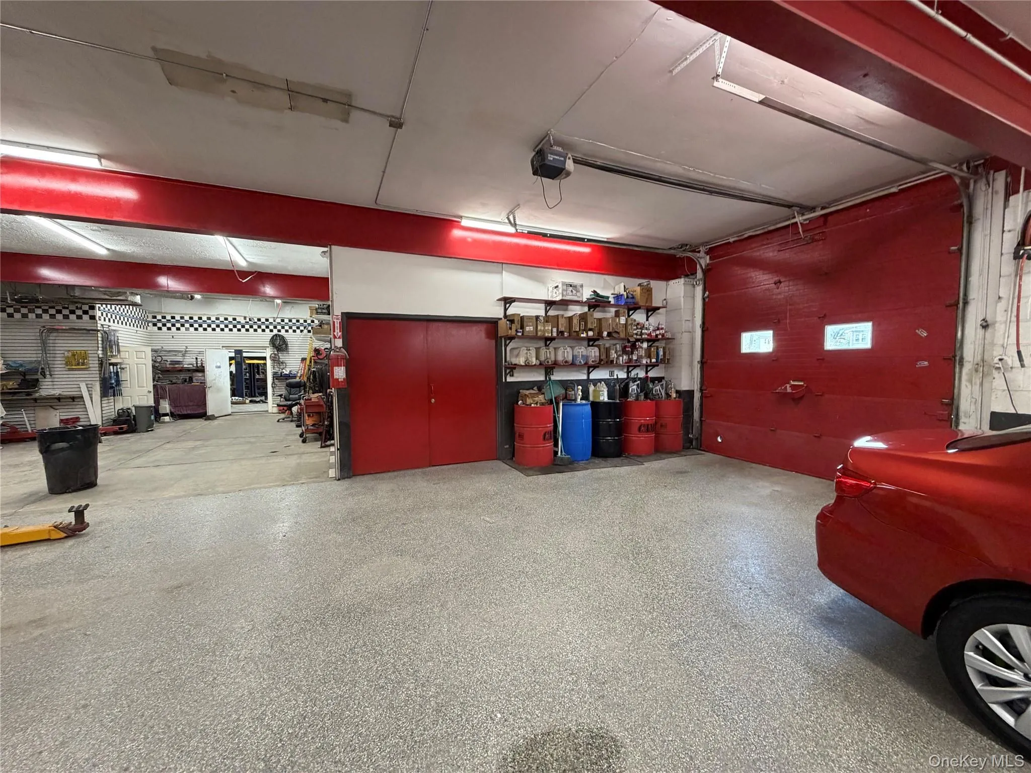 Commercial garage space featuring a red sectional overhead door, built-in shelving, and exposed ceiling beams Commercial garage space featuring a red sectional overhead door, built-in shelving, and exposed ceiling beams