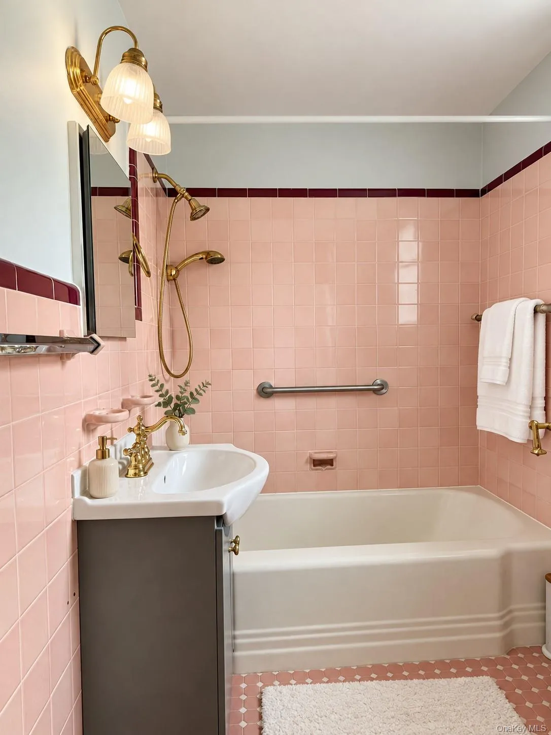Bathroom featuring ceramic wall tiling with burgundy accent trim, a white porcelain pedestal sink with a brass-finish faucet, a brass-finish handheld shower fixture, and a built-in recessed bathtub Bathroom featuring ceramic wall tiling with burgundy accent trim, a white porcelain pedestal sink with a brass-finish faucet, a brass-finish handheld shower fixture, and a built-in recessed bathtub