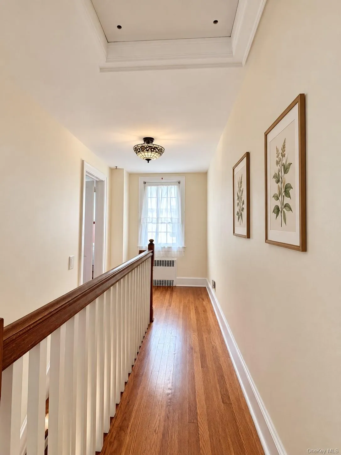 Hallway featuring wood-finish flooring, a decorative glass flush-mount light fixture, a wood-capped handrail with vertical balusters, and a multi-pane window Hallway featuring wood-finish flooring, a decorative glass flush-mount light fixture, a wood-capped handrail with vertical balusters, and a multi-pane window