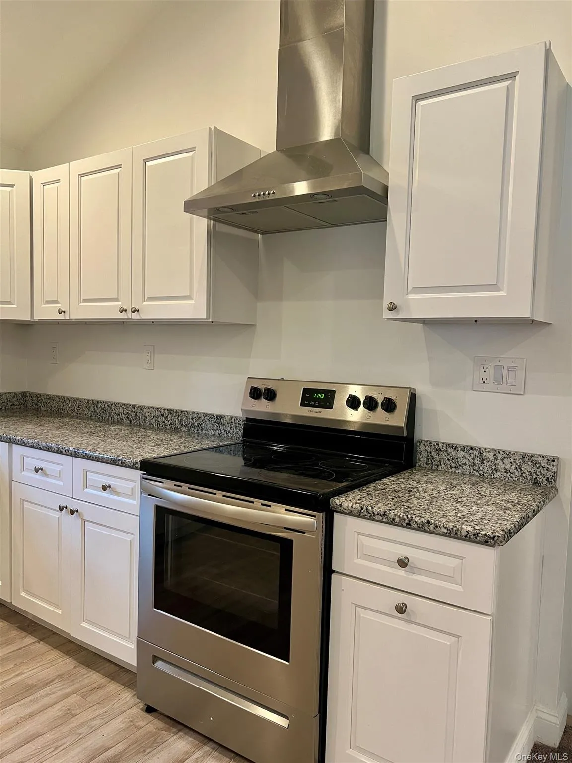Kitchen with stainless steel electric stove, wall chimney exhaust hood, white cabinets, lofted ceiling, and light wood-type flooring Kitchen with stainless steel electric stove, wall chimney exhaust hood, white cabinets, lofted ceiling, and light wood-type flooring