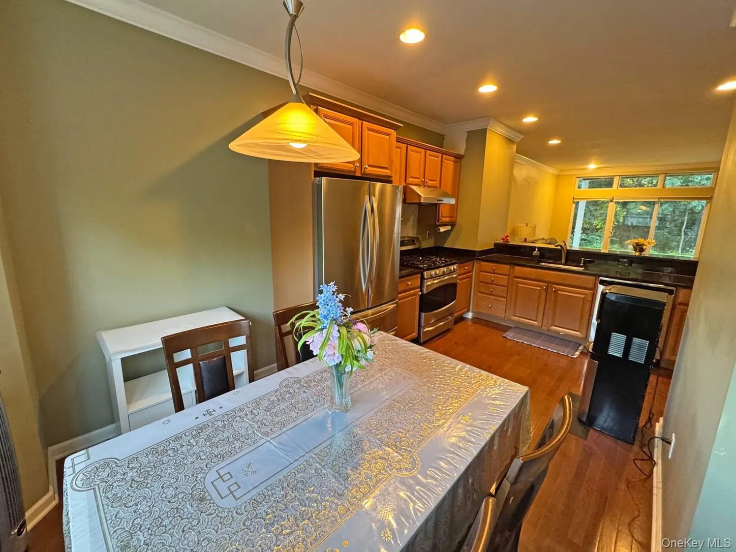 Warm and inviting kitchen and dining area featuring warm wood-finish flooring throughout, stainless steel appliances, custom cabinetry with rich wood tones, granite countertops, and abundant natural light from multiple windows Warm and inviting kitchen and dining area featuring warm wood-finish flooring throughout, stainless steel appliances, custom cabinetry with rich wood tones, granite countertops, and abundant natural light from multiple windows