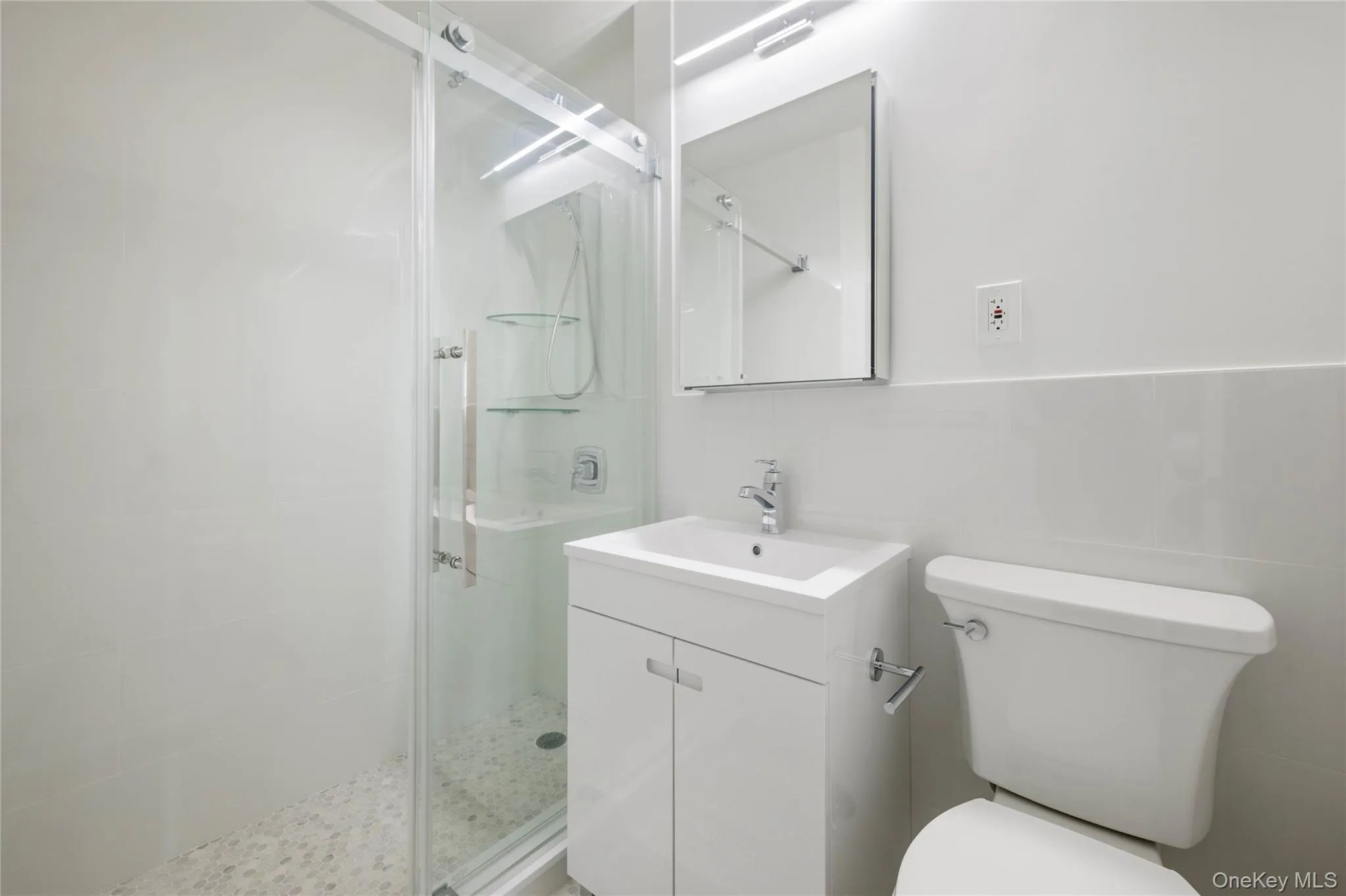 Frameless glass shower enclosure with sliding door, white tile walls, pebble shower floor, vanity with integrated sink, and wall-mounted mirror with overhead lighting Frameless glass shower enclosure with sliding door, white tile walls, pebble shower floor, vanity with integrated sink, and wall-mounted mirror with overhead lighting