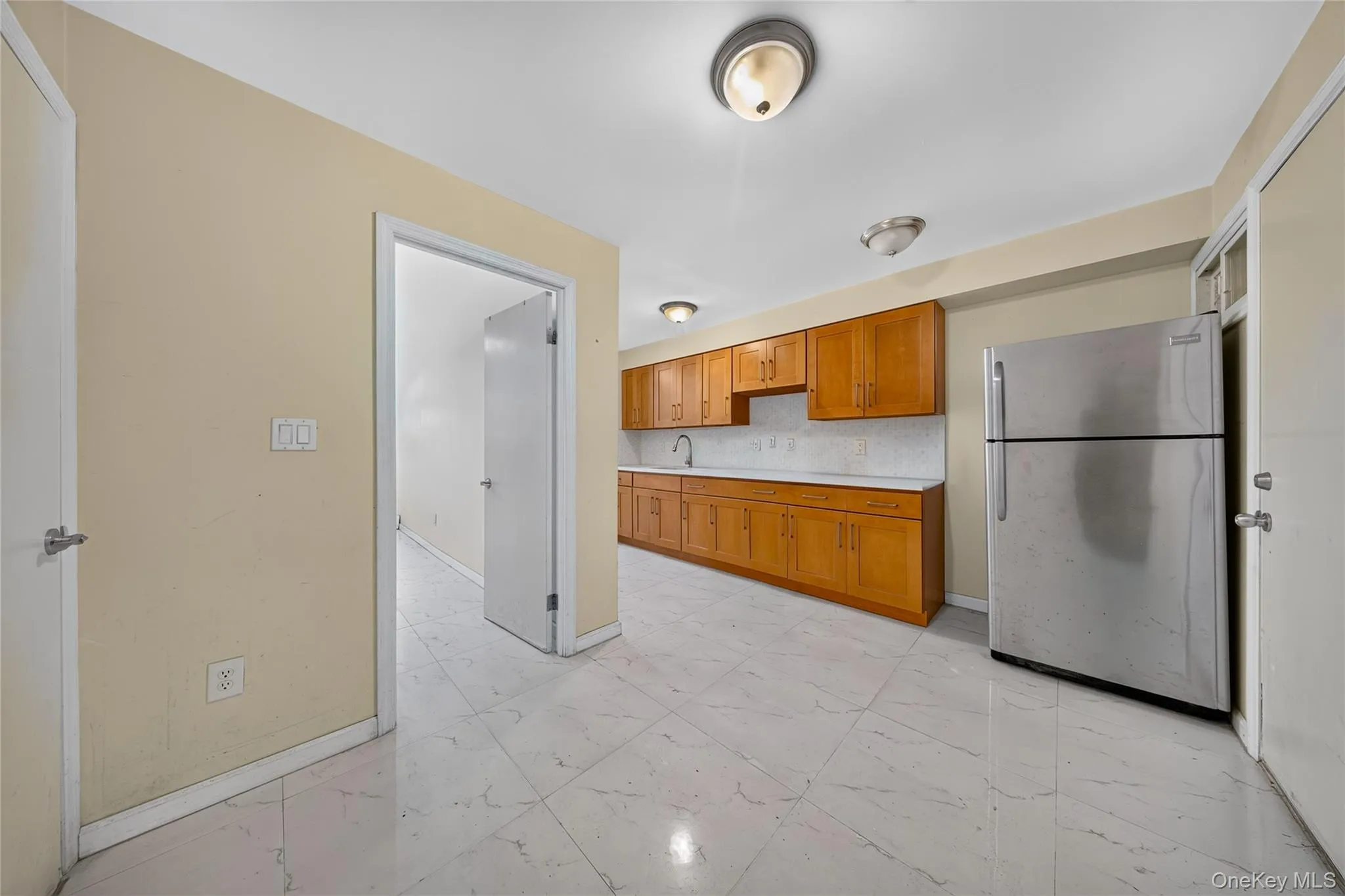 Kitchen area featuring tile flooring, wood-finish cabinetry, stainless steel refrigerator, white countertops, and a white tile backsplash Kitchen area featuring tile flooring, wood-finish cabinetry, stainless steel refrigerator, white countertops, and a white tile backsplash
