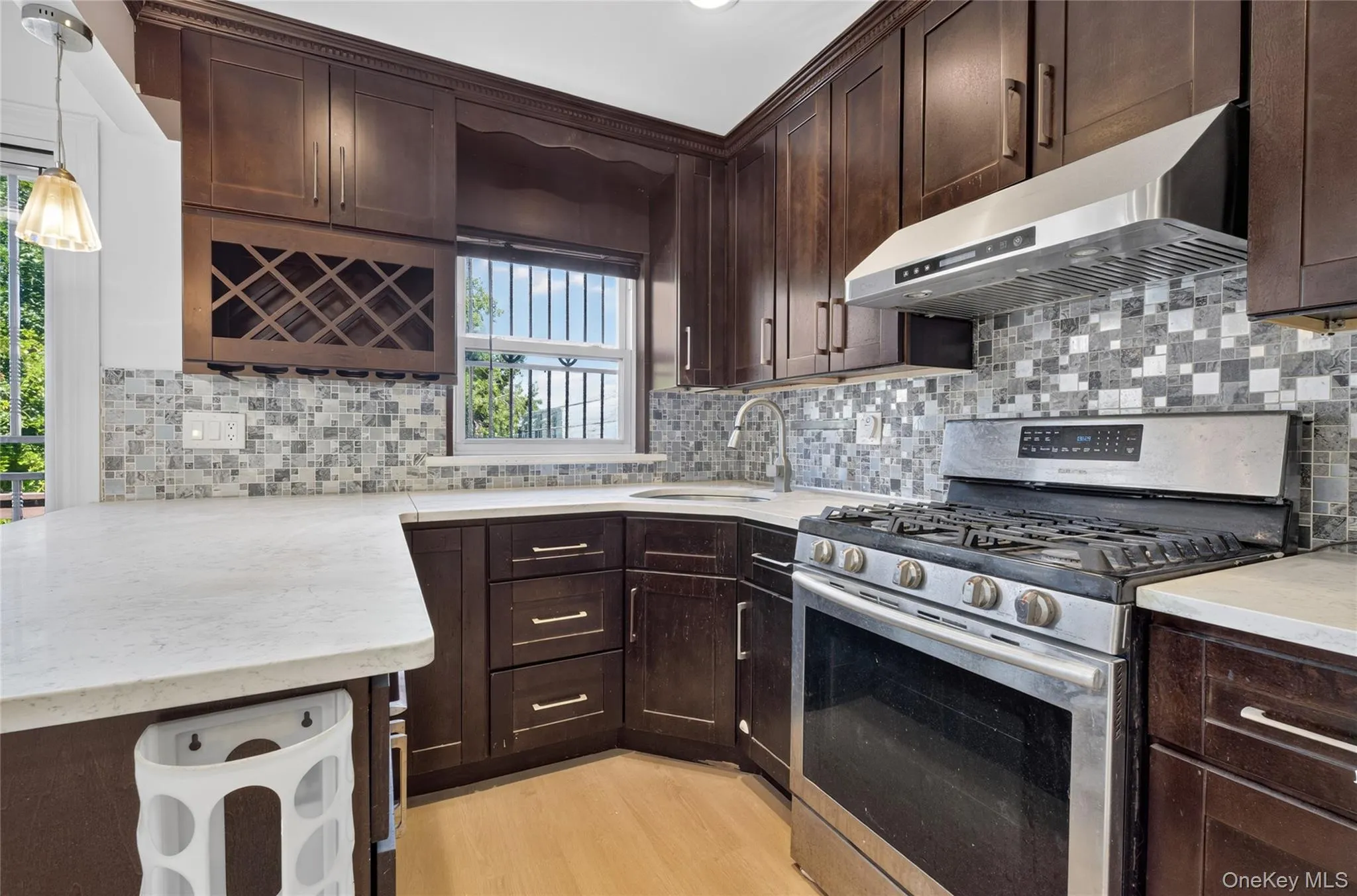 Dark wood-finish cabinetry, mosaic tile backsplash, stainless steel range with overhead hood, undermount sink with a gooseneck faucet, and light-toned countertops Dark wood-finish cabinetry, mosaic tile backsplash, stainless steel range with overhead hood, undermount sink with a gooseneck faucet, and light-toned countertops