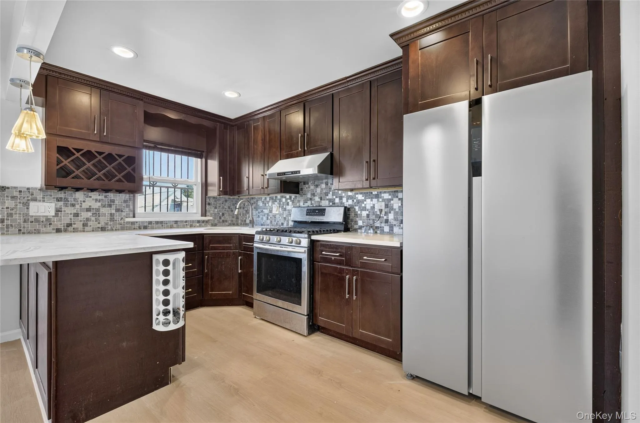 Kitchen featuring dark wood-finish cabinetry, stainless steel appliances, mosaic tile backsplash, light-toned countertops, and wood-finish flooring Kitchen featuring dark wood-finish cabinetry, stainless steel appliances, mosaic tile backsplash, light-toned countertops, and wood-finish flooring