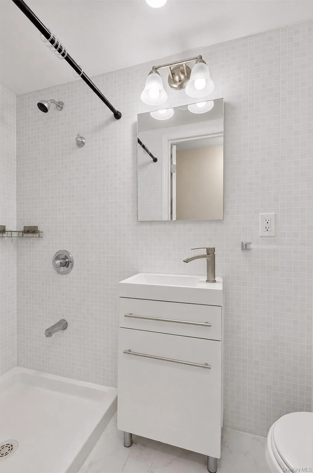 White square tile walls, built-in shower, freestanding vanity with integrated sink, rectangular wall mirror, and two-light vanity fixture White square tile walls, built-in shower, freestanding vanity with integrated sink, rectangular wall mirror, and two-light vanity fixture