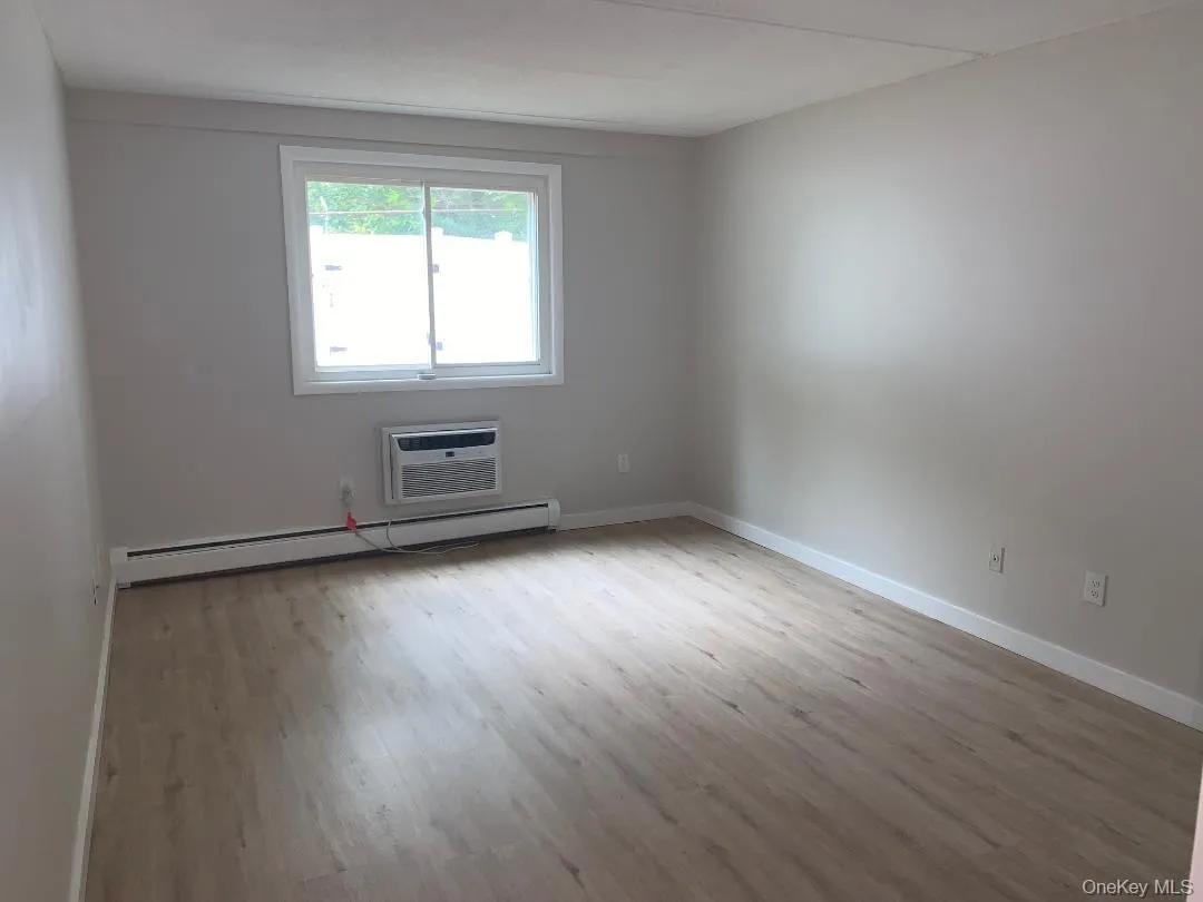 Spacious primary bedroom with light wood-style floors, a baseboard heating unit, and a wall mounted air conditioner Spacious primary bedroom with light wood-style floors, a baseboard heating unit, and a wall mounted air conditioner