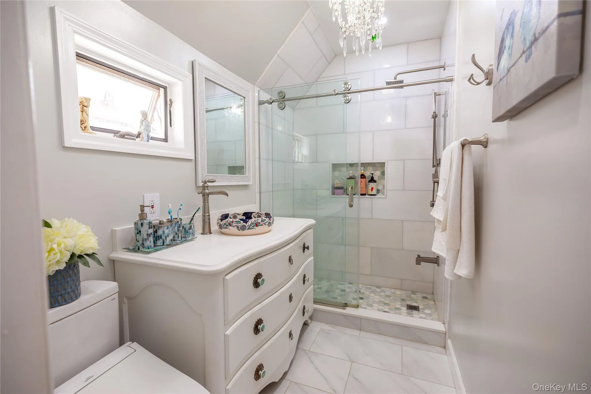 Spa-inspired primary bathroom featuring a custom white vanity with multiple drawers, white subway tile surround, and a frameless glass walk-in shower with a rainfall showerhead and built-in shelving Spa-inspired primary bathroom featuring a custom white vanity with multiple drawers, white subway tile surround, and a frameless glass walk-in shower with a rainfall showerhead and built-in shelving