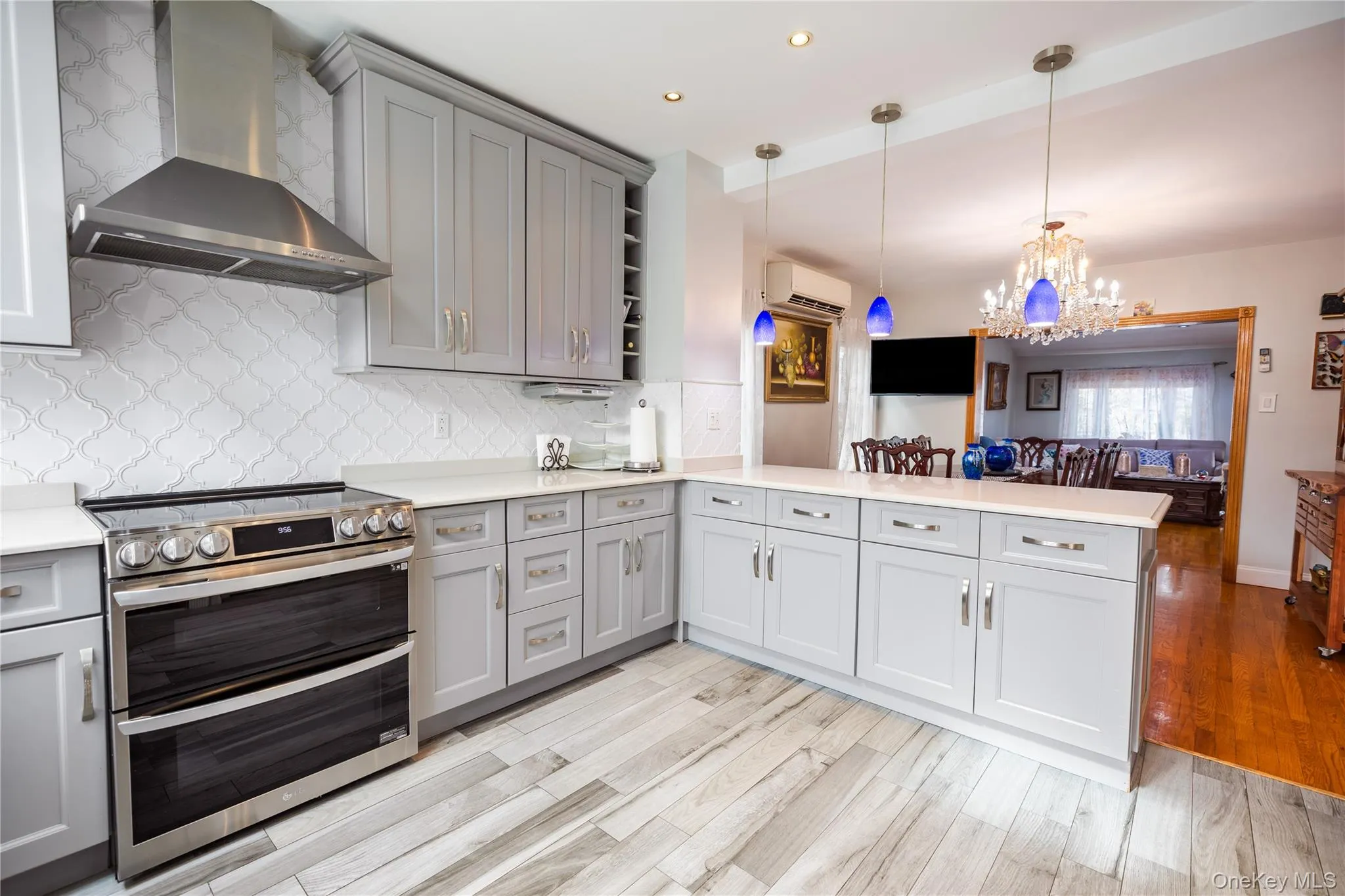 Light gray shaker cabinetry with brushed nickel hardware, light-toned countertops, stainless steel range with matching hood, patterned tile backsplash, and wood-finish flooring Light gray shaker cabinetry with brushed nickel hardware, light-toned countertops, stainless steel range with matching hood, patterned tile backsplash, and wood-finish flooring