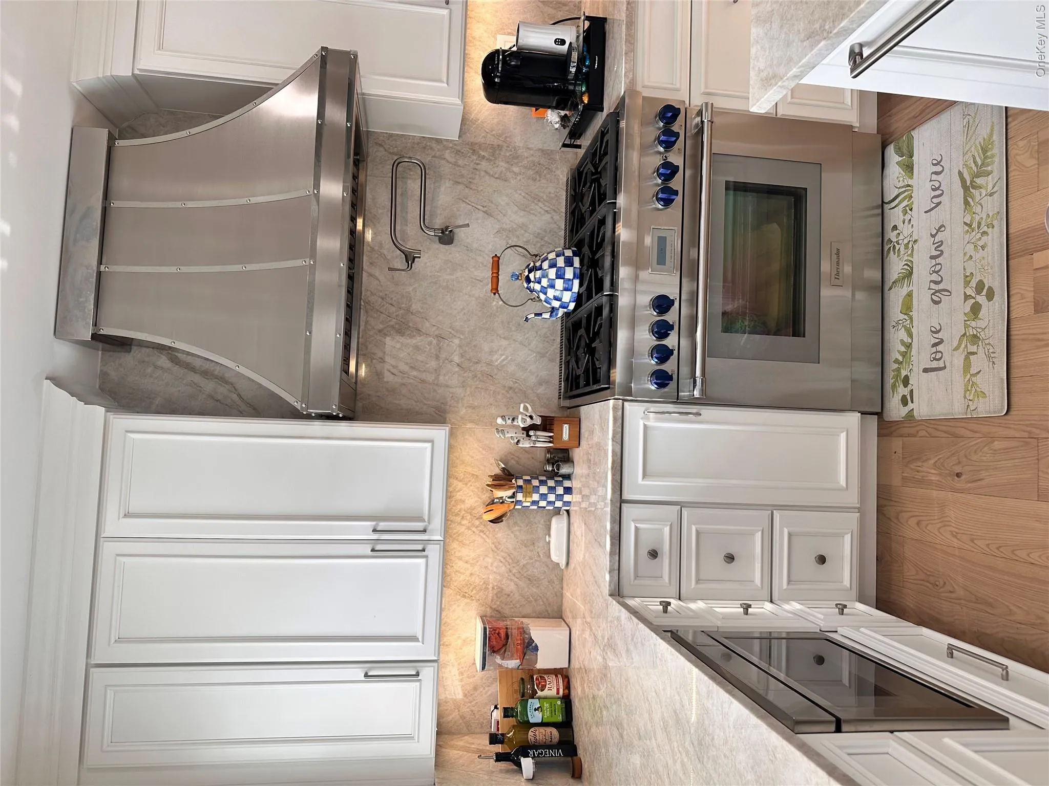 White shaker-style cabinetry, stainless steel range hood, built-in microwave drawer, light-toned stone countertops, and wood-finish flooring White shaker-style cabinetry, stainless steel range hood, built-in microwave drawer, light-toned stone countertops, and wood-finish flooring