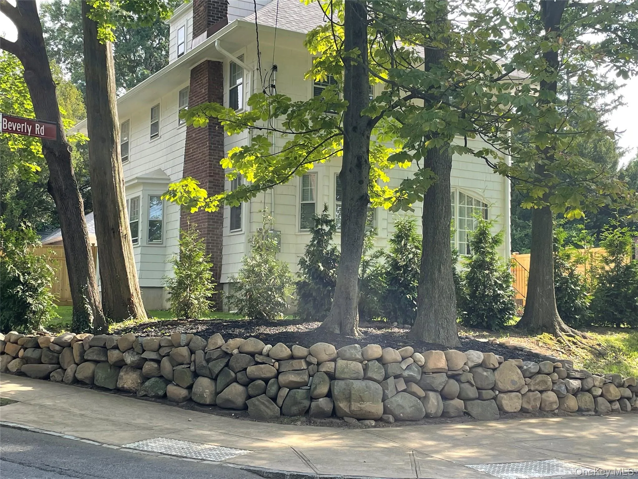 White exterior siding, stone retaining wall, mature trees, corner lot, bay window White exterior siding, stone retaining wall, mature trees, corner lot, bay window