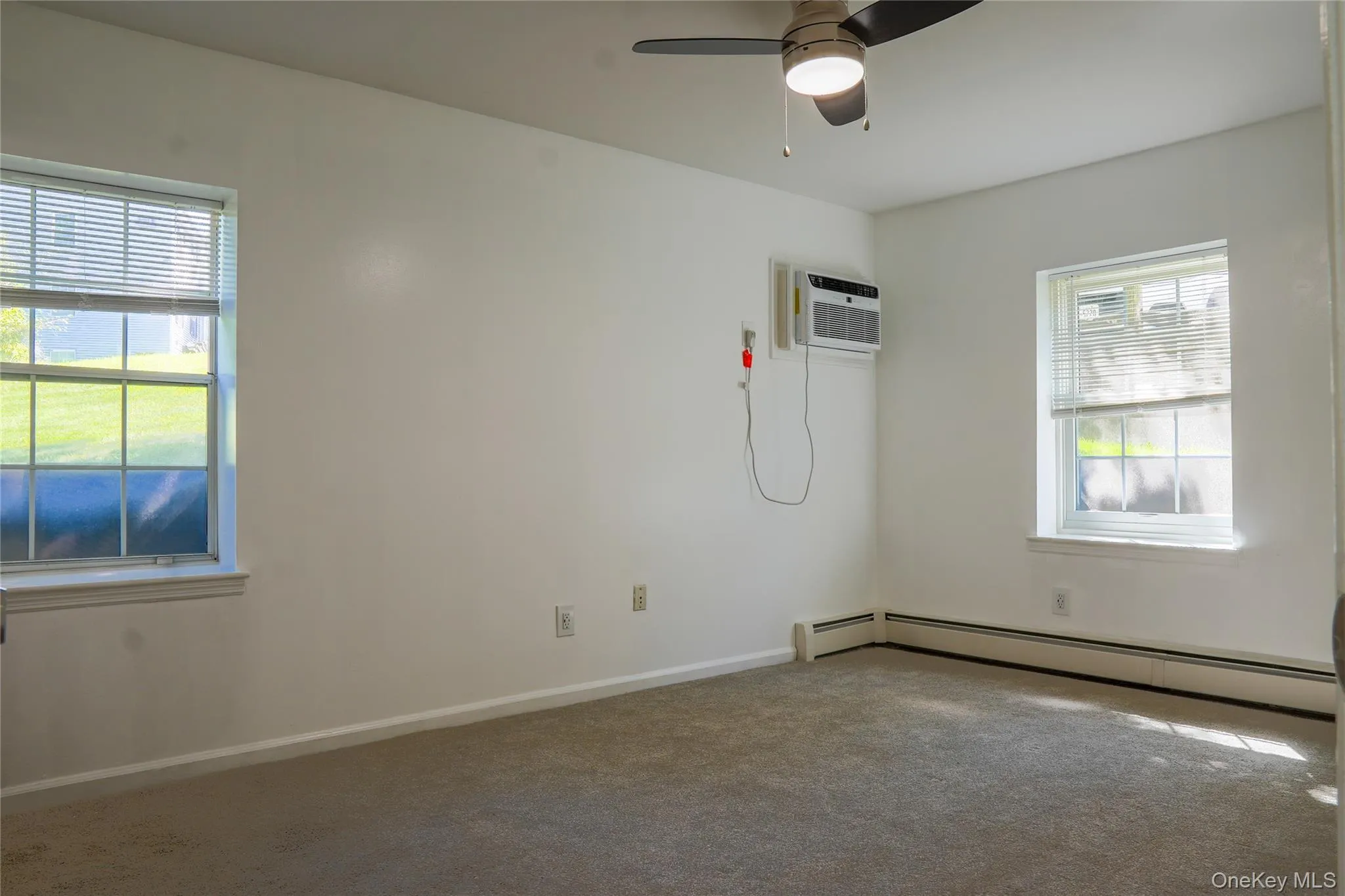 Primary bedroom with carpeted flooring, white walls, two windows with blinds, ceiling fan with integrated lighting, and a wall-mounted air conditioning unit Primary bedroom with carpeted flooring, white walls, two windows with blinds, ceiling fan with integrated lighting, and a wall-mounted air conditioning unit