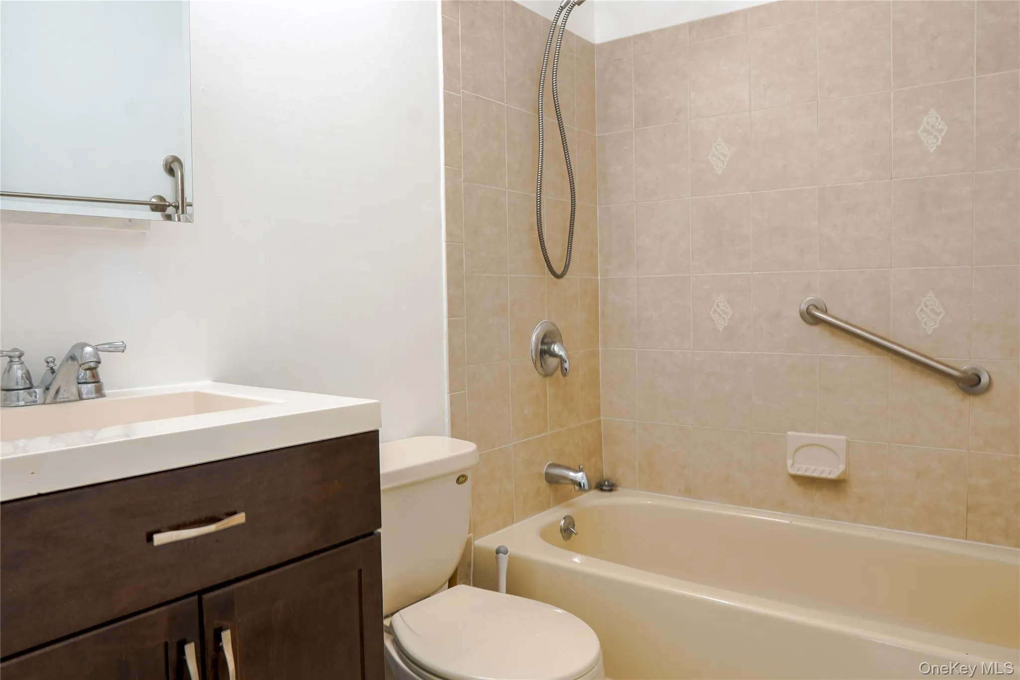 Primary bathroom featuring a dark wood-finish vanity, white countertop with integrated sink, chrome faucet, and tiled shower with grab bar Primary bathroom featuring a dark wood-finish vanity, white countertop with integrated sink, chrome faucet, and tiled shower with grab bar