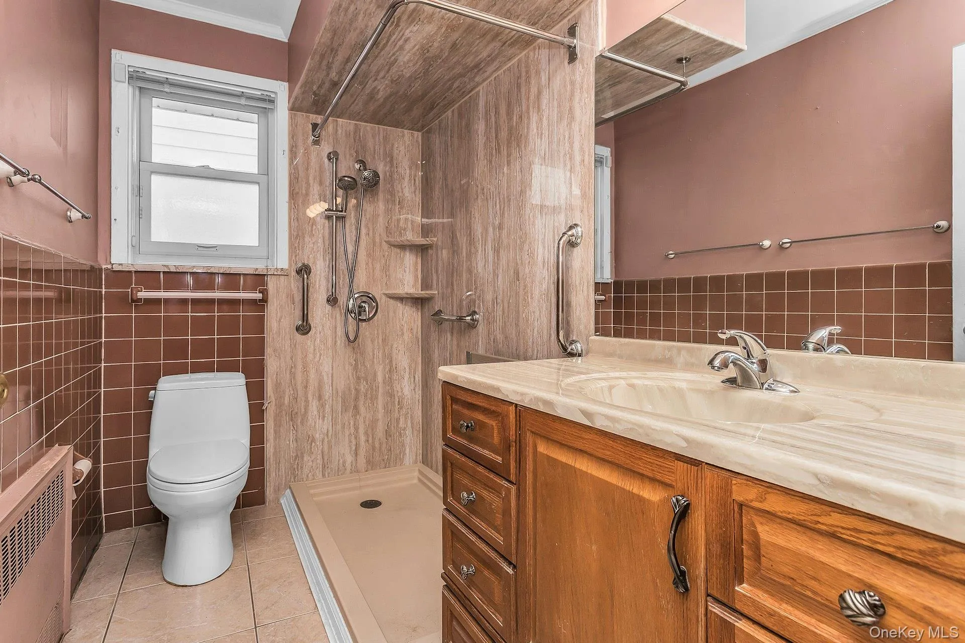 Bathroom featuring a wood-finish vanity with an integrated sink, a large mirror, a tiled shower with grab bars, and a window Bathroom featuring a wood-finish vanity with an integrated sink, a large mirror, a tiled shower with grab bars, and a window