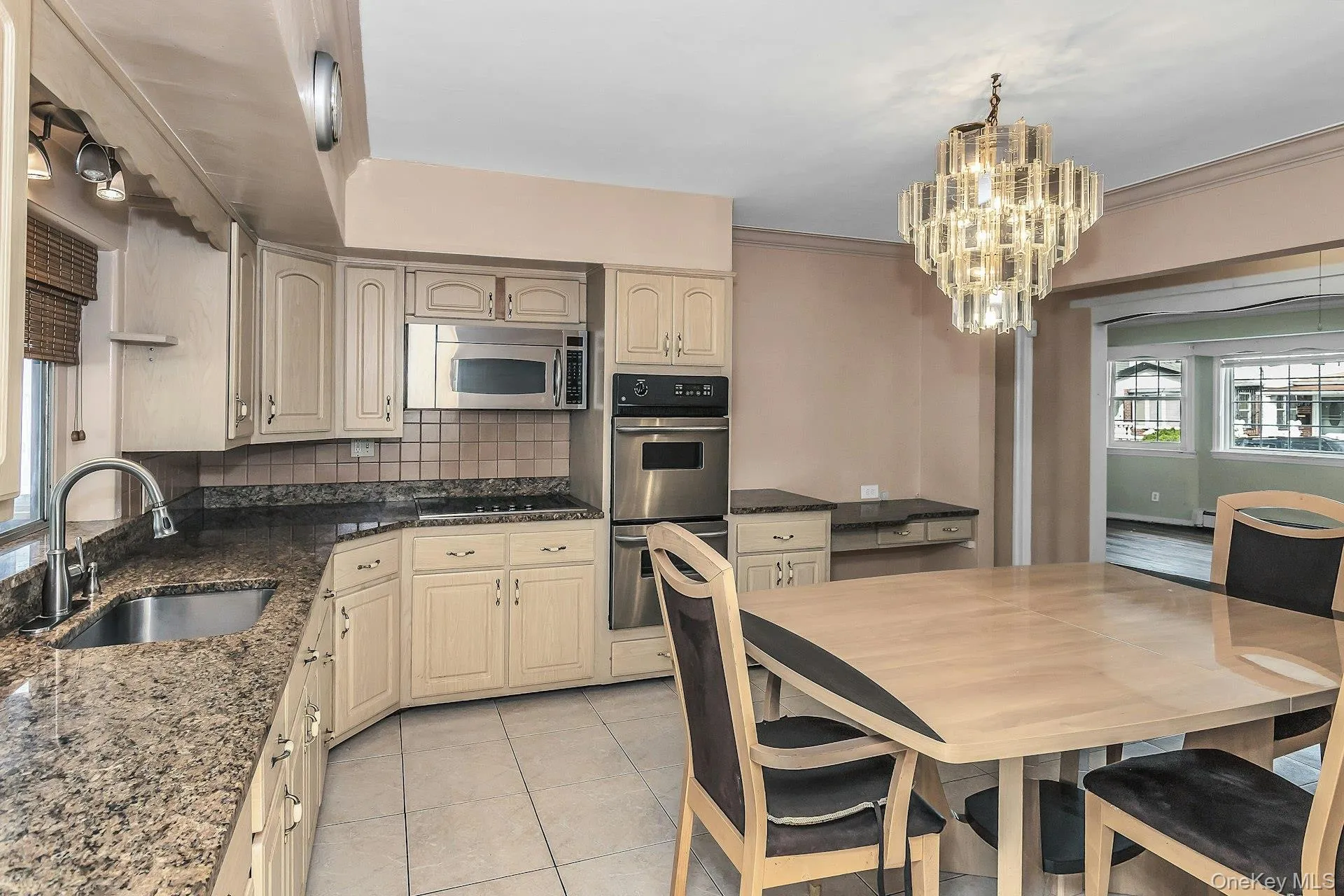 Kitchen featuring light-toned cabinetry, granite-finish countertops, stainless steel appliances, a tile backsplash, and a contemporary chandelier Kitchen featuring light-toned cabinetry, granite-finish countertops, stainless steel appliances, a tile backsplash, and a contemporary chandelier