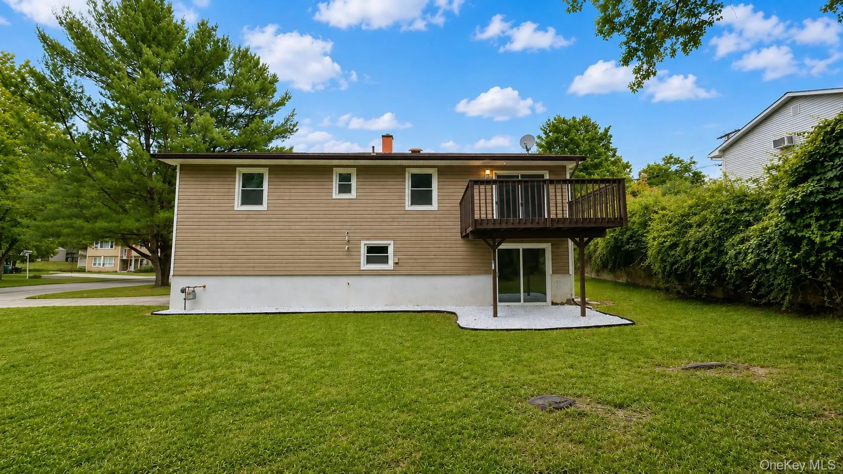 Rear elevation featuring a raised wood deck, ground-level patio with white gravel, and a sliding glass door Rear elevation featuring a raised wood deck, ground-level patio with white gravel, and a sliding glass door